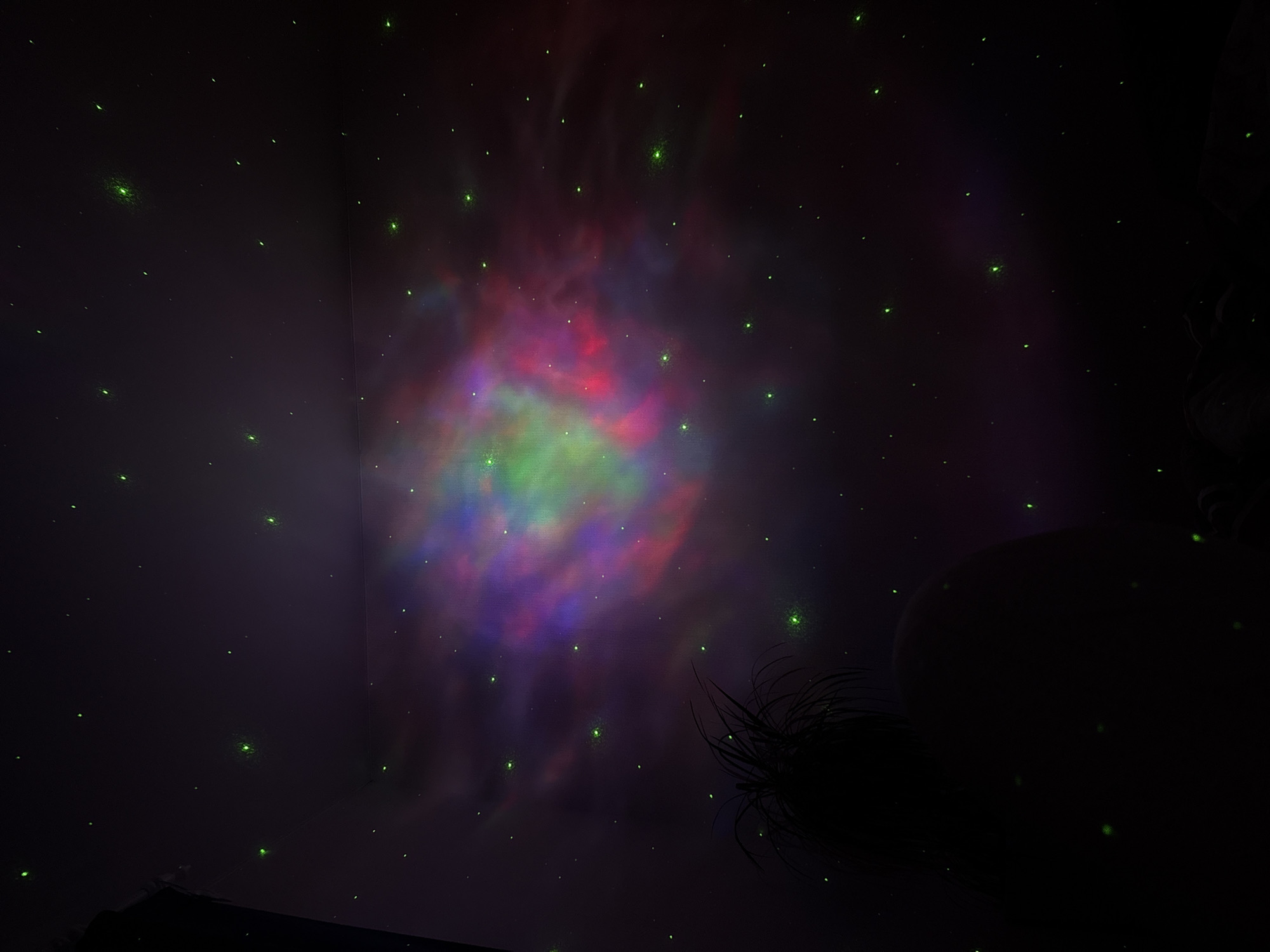 This is a photograph of a bedroom ceiling in the city of Perm, Russia, illuminated by a star projector. The projector casts a nebula-like pattern of colored light, with hues of purple, pink, and green, across the dark ceiling. Numerous small, green dots, representing stars, are scattered throughout the projection. In the foreground, partially silhouetted, is the back of a person's head with spiky hair, and a section of their shoulder or arm. The overall impression is of a dimly lit, intimate space transformed into a cosmic spectacle.