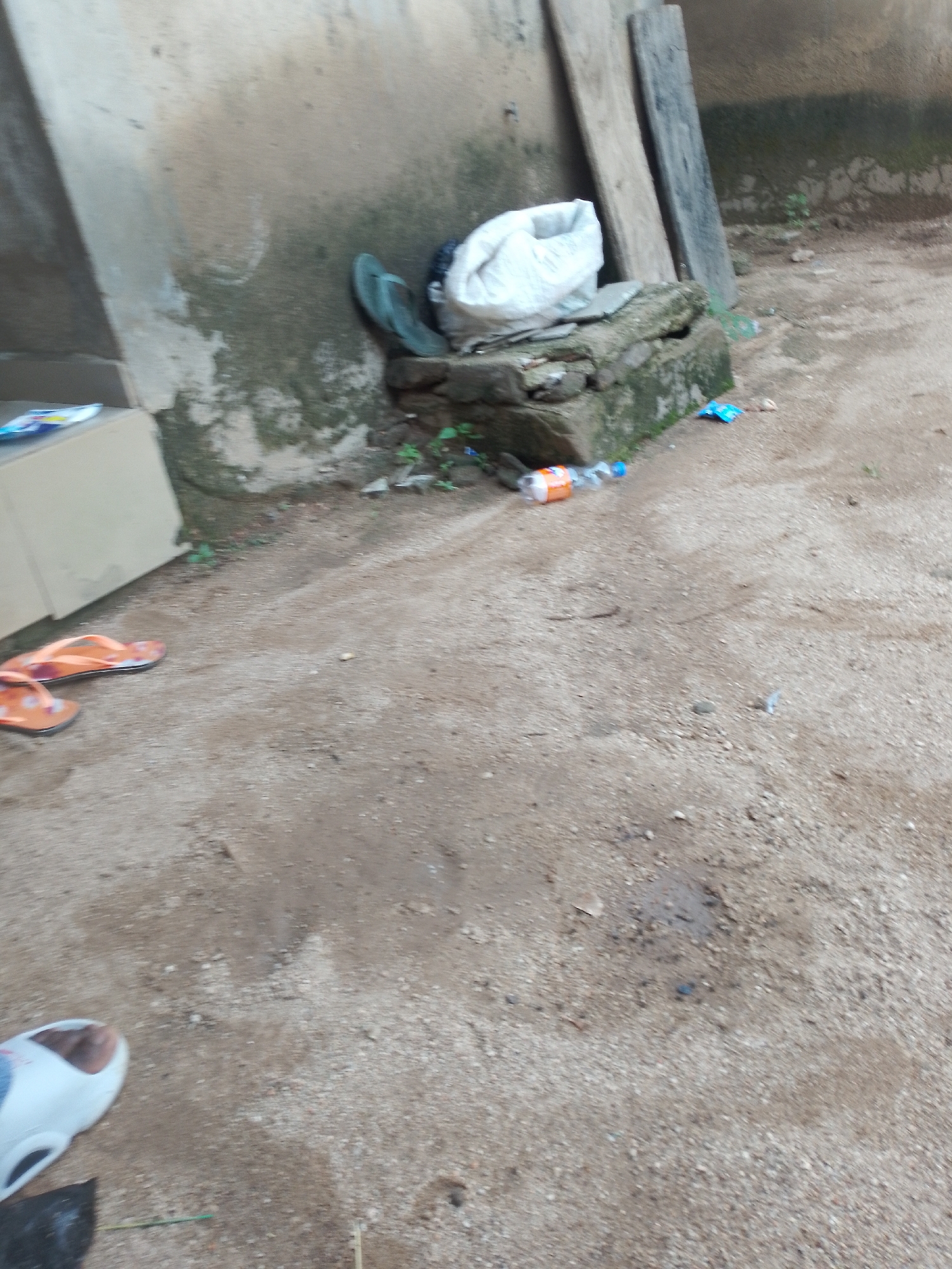 The image shows a ground-level view of an outdoor area with a concrete wall in the background and sandy, gravelly ground in the foreground. Several objects are scattered around, including a pair of orange flip-flops, a white plastic bag, a green flip-flop, and a plastic bottle with an orange label lying on its side. There is also a white shoe on the lower left side of the image. The scene appears to be in a state of disarray, with debris and discarded items.