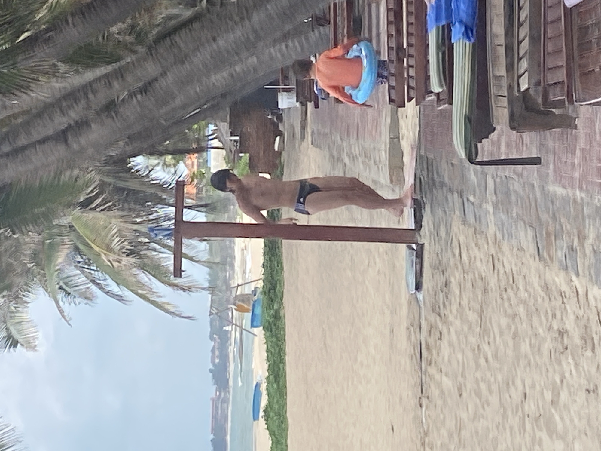 The image depicts a sandy beach scene with palm trees in Phu Thuy Ward, Vietnam. In the foreground, a man in dark swim trunks stands under a shower fixture. To the right, a child wearing an orange shirt and patterned swim trunks is holding a blue inner tube. Rows of wooden lounge chairs with cushions are visible to the right of the paved walkway. In the background, the ocean meets a coastline with buildings and more vegetation. Several blue floatation devices are present on the sand near the water. The sky is overcast.