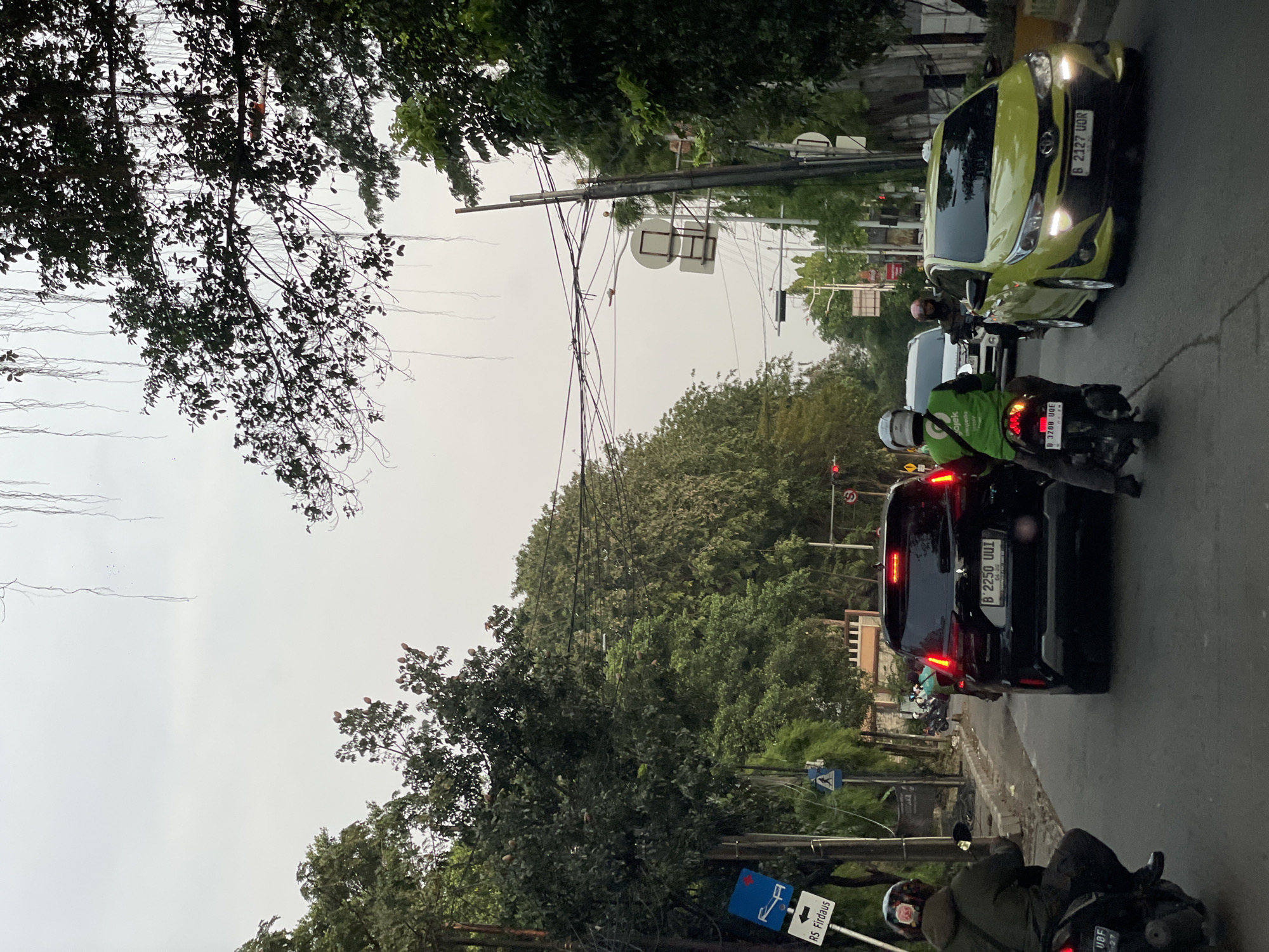 The image depicts a street scene in North Jakarta, Indonesia. A yellow car with license plate B 2327 UQR is stopped at a traffic light. A black car with license plate B 2250 UUI is positioned behind it, with a motorcyclist alongside. Another motorcyclist wearing a green jacket with a Gojek logo is also present. Overhead, there is a dense network of electrical wires amidst tree branches. There are signs that read “RS Firdaus”. The sky is overcast.