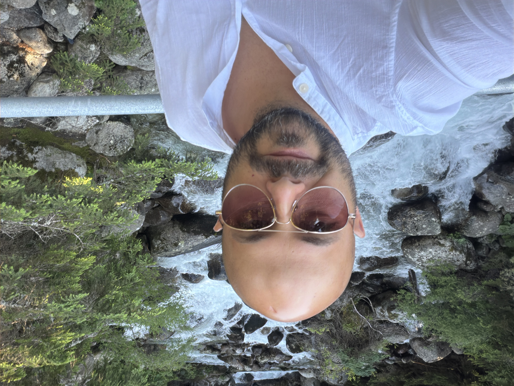 A man with a shaved head, wearing aviator sunglasses and a white button-down shirt, takes a selfie in front of a rushing mountain stream with rocky banks and evergreen trees. The man's face is centrally located in the frame, and his body extends to the right. The stream is the primary background element, with whitewater cascading over rocks. Lush green foliage and large boulders line the stream's edges. A metal railing runs horizontally across the lower right portion of the image. The setting appears to be a natural, outdoor environment, possibly a hiking trail or scenic viewpoint. The image quality is good, with clear detail and natural lighting. The location is identified as Bansko, Bulgaria.
