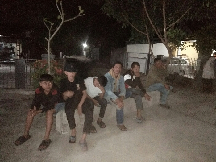 The image displays six males seated outdoors at night in what appears to be a residential street or communal area in Baleendah, Indonesia. They are positioned along a low, concrete wall or bench. The scene is dimly lit, indicating it is nighttime, with ambient artificial light sources providing illumination.
From left to right, the subjects include a young boy in a black shirt with red accents and sandals, notable for red-eye from the camera flash. Beside him is a male wearing a black cap, black t-shirt, black pants, and sandals. The third individual wears a white t-shirt, light blue jeans, and sandals, leaning forward with his head down. Next is a man in a light blue denim jacket over a checkered shirt, dark pants, and sandals. The fifth subject, with a beard, is dressed in a dark jacket and dark pants, looking away from the camera. The rightmost individual wears a light brown or olive jacket, blue jeans, and dark shoes, also looking away.
In the background, trees with varying foliage density are visible, along with shrubs featuring red flowers. Beyond these, elements of a residential street can be seen, including houses, fences, and a white car parked behind a gate. Faint lights in the distance suggest streetlights or lights from buildings. The group appears to be casually gathered, sitting together, possibly socializing or resting. No specific event or intense activity is evident. No text is visible in the image. The overall impression is a relaxed, informal gathering during the evening hours in a local neighborhood setting.