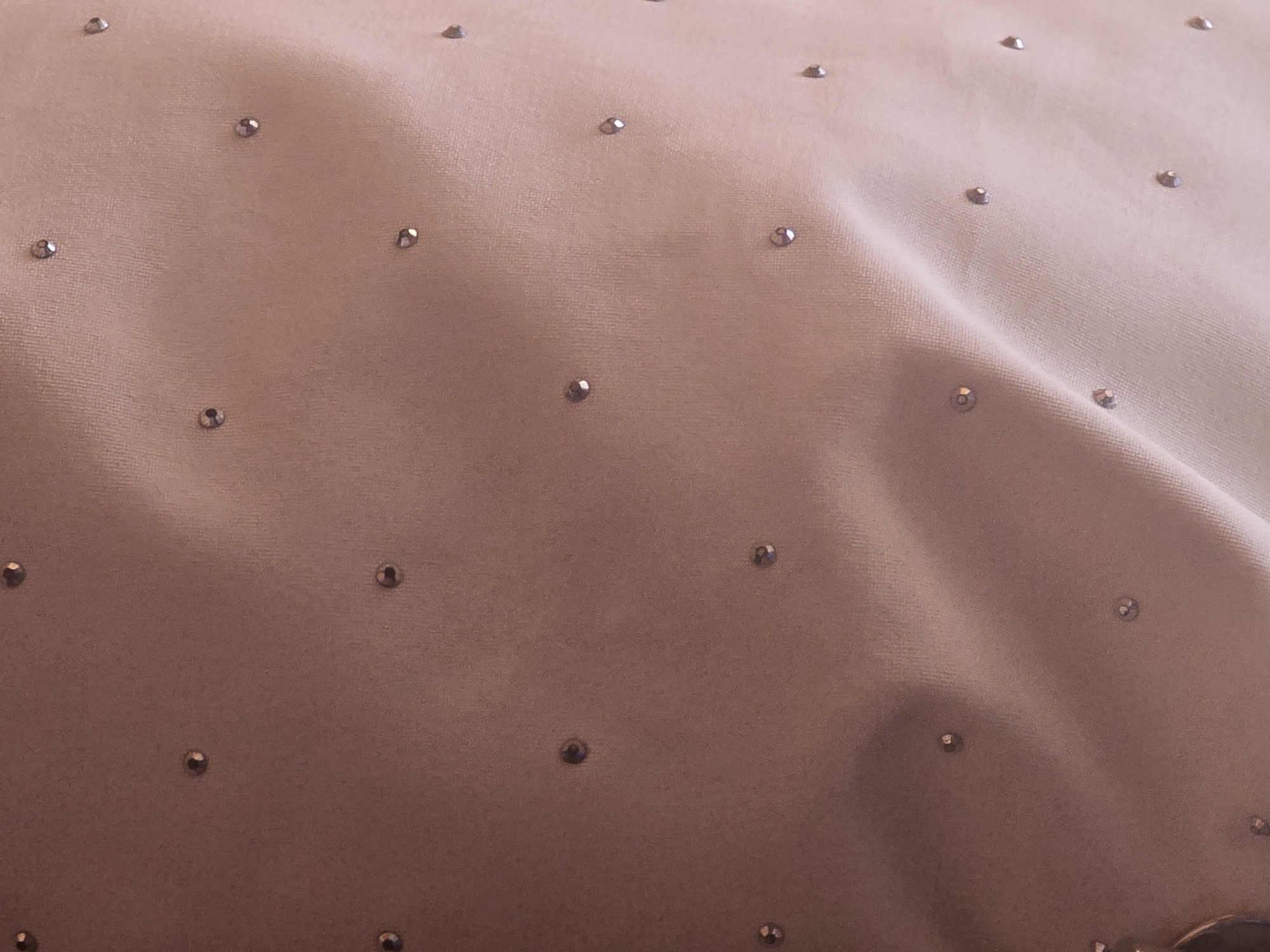 The image shows a close-up view of a fabric surface with a repeating pattern of small, reflective embellishments. The fabric appears to be a light, neutral color, possibly beige or light pink, with a soft texture. The embellishments are scattered across the surface, creating a shimmering effect. There are no people, specific landmarks, or discernible environmental features visible in the image that would allow for identification of a location in Maksimiha, Russia. The focus is solely on the texture and decorative elements of the material.