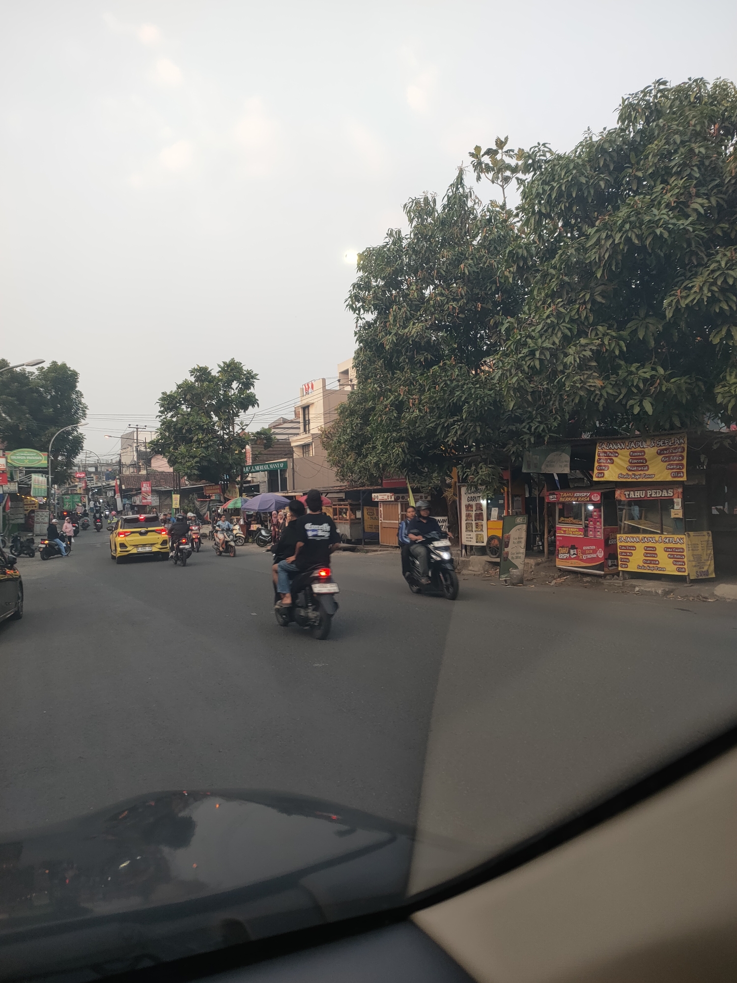 The image captures a bustling street scene in Cimahi, Indonesia, likely during the daytime. A busy road with multiple lanes is filled with various vehicles, predominantly motorcycles, which are a common mode of transportation in the region. Several motorcycles are seen carrying passengers, indicating active daily commuting. A bright yellow car is also visible in the traffic. Along the sides of the road, numerous small shops and food stalls are set up, with some displaying colorful signage and menus. Trees line the street, providing some greenery amidst the urban environment. Buildings of varying sizes and architectural styles are present, suggesting a mixed commercial and residential area. The sky appears overcast, contributing to a diffused lighting condition. The overall impression is that of a lively urban setting characterized by active commerce and daily life.