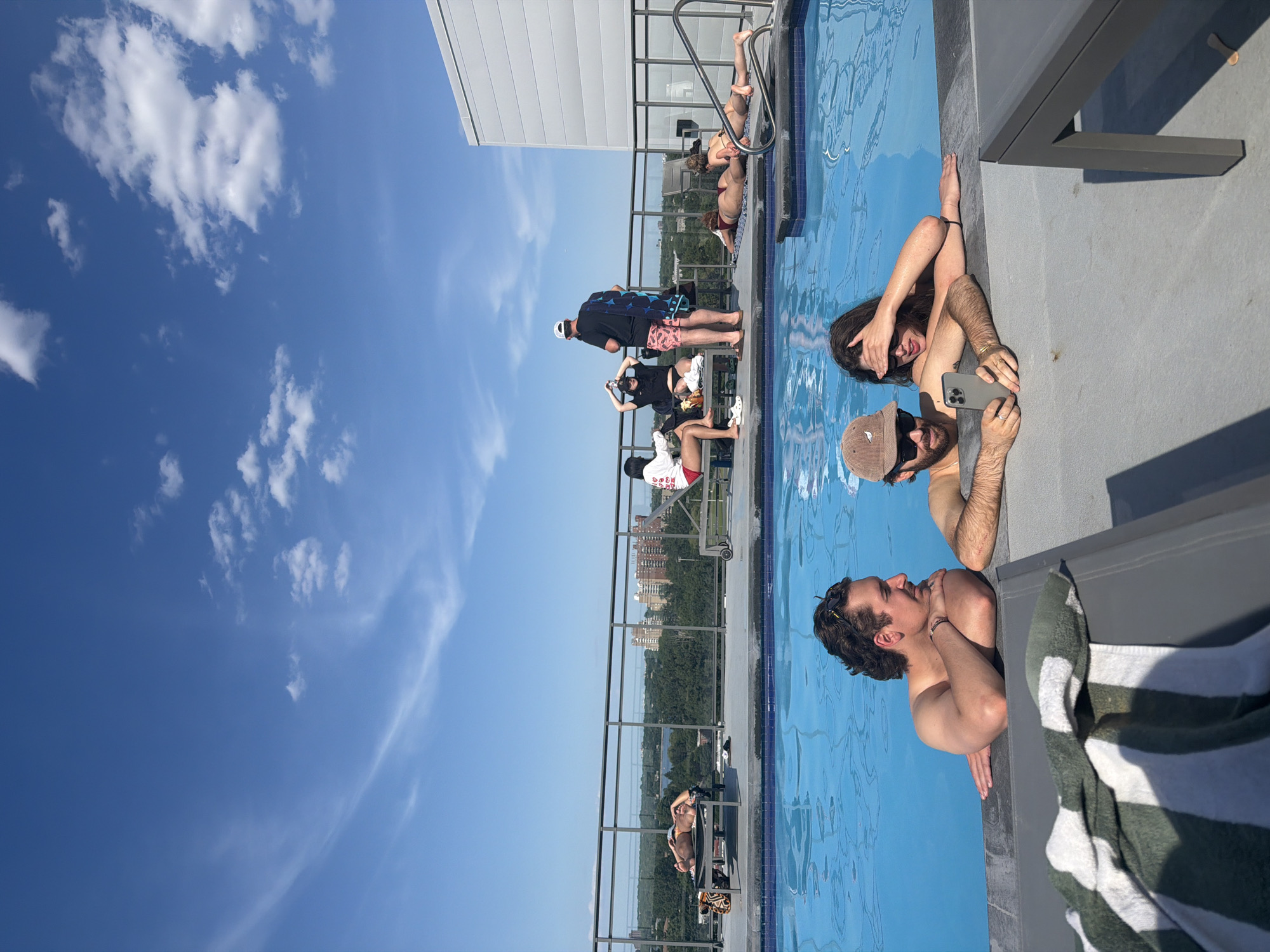 The image depicts a sunny day at a rooftop swimming pool, possibly in Laval, Canada, with several people relaxing. In the foreground, two individuals are partially submerged in the pool. One, a man with dark curly hair and a mustache, leans on the pool edge, looking to his right. Next to him, a man wearing a beige baseball cap and sunglasses is taking a selfie with a smartphone, his arm around a woman who is also in the pool, shielding her eyes from the sun.
In the background, several other individuals are scattered around the pool area. Some are lounging on chairs and towels on the deck, soaking up the sun, while others appear to be standing and conversing. Beyond the pool and deck, a cityscape is visible, with buildings and trees indicating an urban or suburban environment. The sky is clear blue with scattered white clouds, and the overall atmosphere suggests a leisurely, recreational setting.