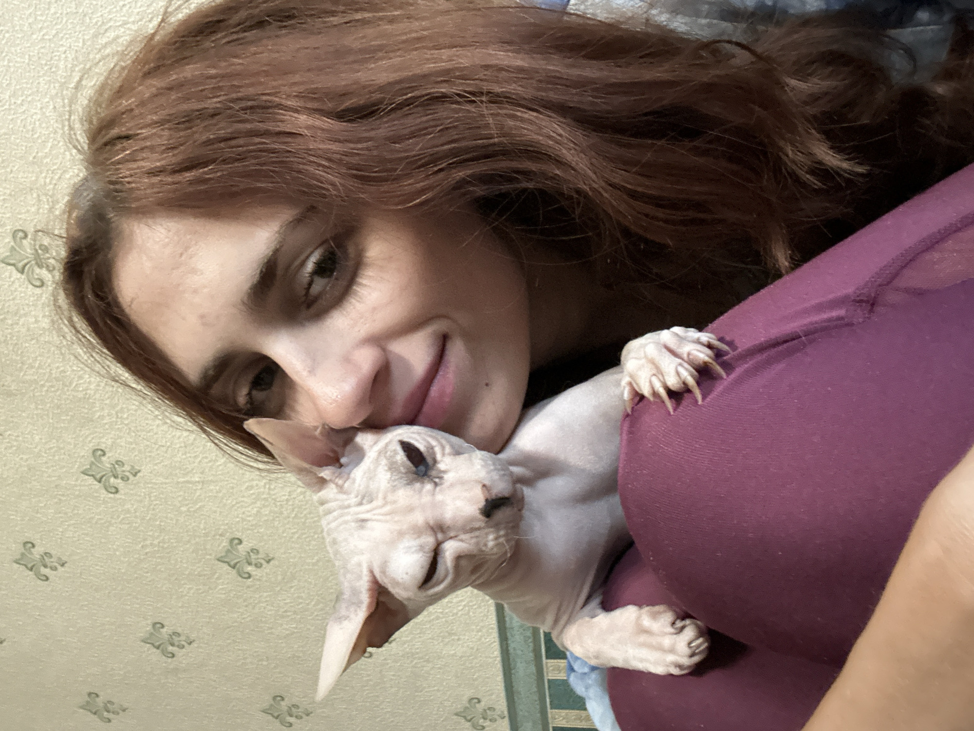 A young woman with long, reddish-brown hair and brown eyes is holding a light-skinned Sphynx cat. The cat is positioned on her lap, leaning its head against the woman's cheek, and has its eyes partially closed, appearing to squint. The woman is smiling gently. She is wearing dark purple leggings. The background shows a textured wallpaper with a repeating floral pattern in a light, neutral color. The scene is an indoor environment.