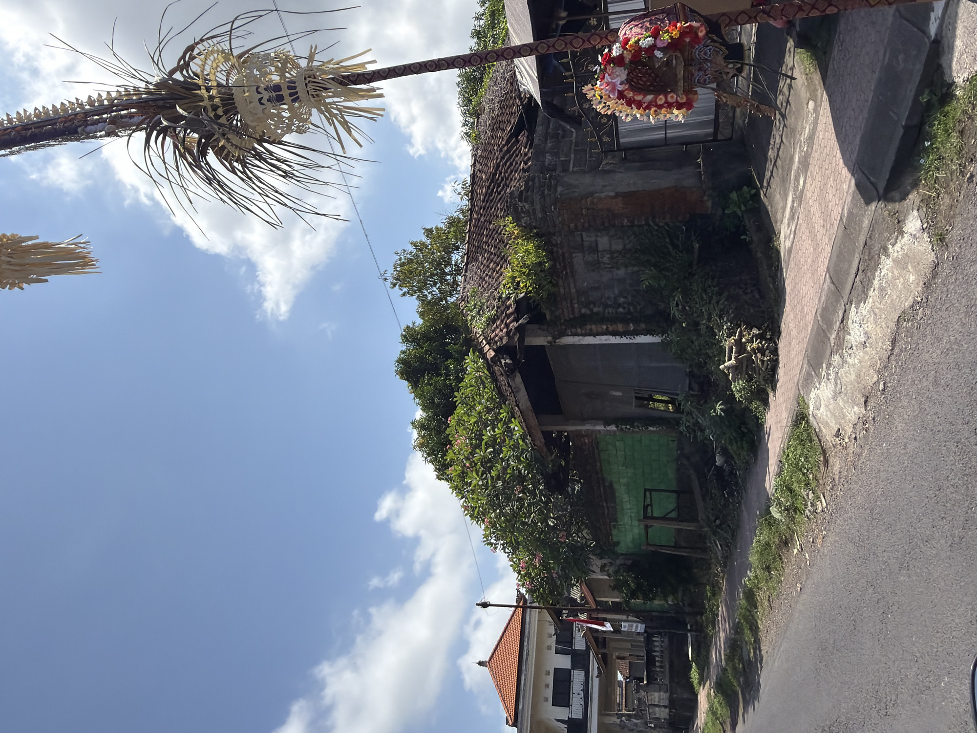 This is a street-level, medium-angle, full-shot photograph of a residential area in Blahbatuh, Indonesia, on a bright, cloudy day. The foreground features an asphalt road with a paved sidewalk running alongside it. The sidewalk is bordered by a concrete curb and overgrown grass. To the right of the sidewalk, a tall, elaborately decorated pole stands, adorned with woven palm leaves and what appears to be a floral arrangement at its base.
Behind the sidewalk, there are several structures. A building with a partially collapsed, red-tiled roof is prominent, with brick walls and green-painted sections. Vegetation, including trees and shrubs, is growing over and around this building. To the left of this structure, a more modern two-story building with white walls and a red-tiled roof is visible, featuring balconies and windows. A small Indonesian flag is attached to a pole in front of this building. The overall scene suggests a mix of older, possibly decaying buildings alongside more contemporary residences, set against a backdrop of lush greenery and a blue sky with scattered clouds. There are no people visible in the photograph.