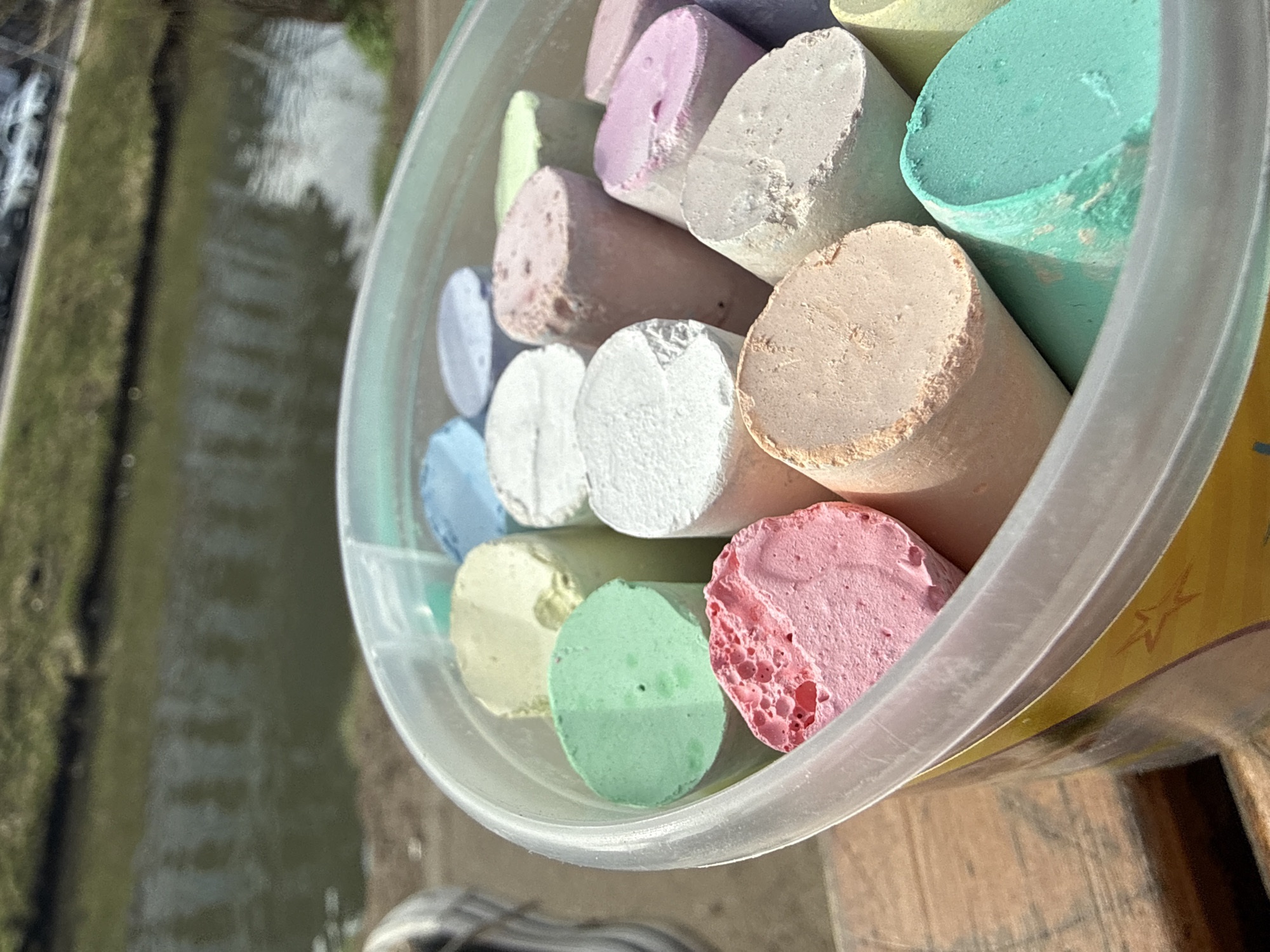 The image shows a clear plastic bucket filled with pieces of colorful sidewalk chalk. The chalk is in cylindrical shapes and includes colors like pink, white, blue, green, yellow, and lavender. The bucket sits on a wooden surface, potentially a table or deck. In the background, partially visible, is a blurred view of a waterway bordered by green vegetation. There is no evidence to support this being in the city of Dmitrov, Russia.