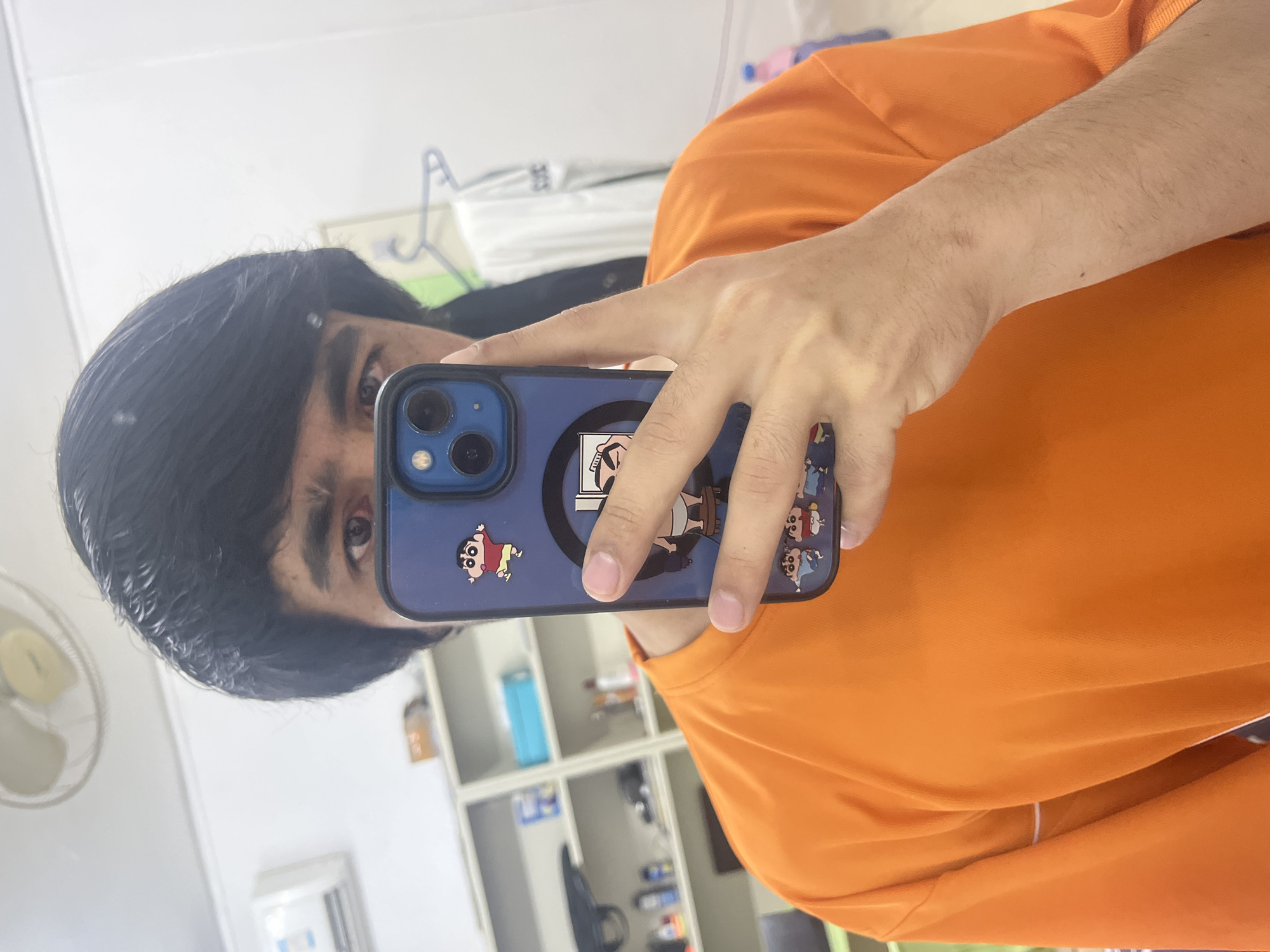 An indoor mirror selfie captures a person holding a blue smartphone. The person's face is partially visible, showing dark hair and eyes, and they are wearing a bright orange t-shirt. Their right hand (appearing as the left due to the mirror reflection) holds the phone, which has a blue case decorated with multiple cartoon characters, including what appears to be Crayon Shin-chan.
The background, reflected in the mirror, shows a room with white walls. Visible elements include a white shelf unit containing various objects like bottles and boxes, a partially obscured white fan in the upper left, and a white item possibly hanging on a hook in the upper background. The setting suggests a casual interior space, possibly an office or living area. No specific time of day is indicated, with the scene lit by indoor lighting. The image is rotated 90 degrees clockwise. No discernible text is visible other than the cartoon characters on the phone case.