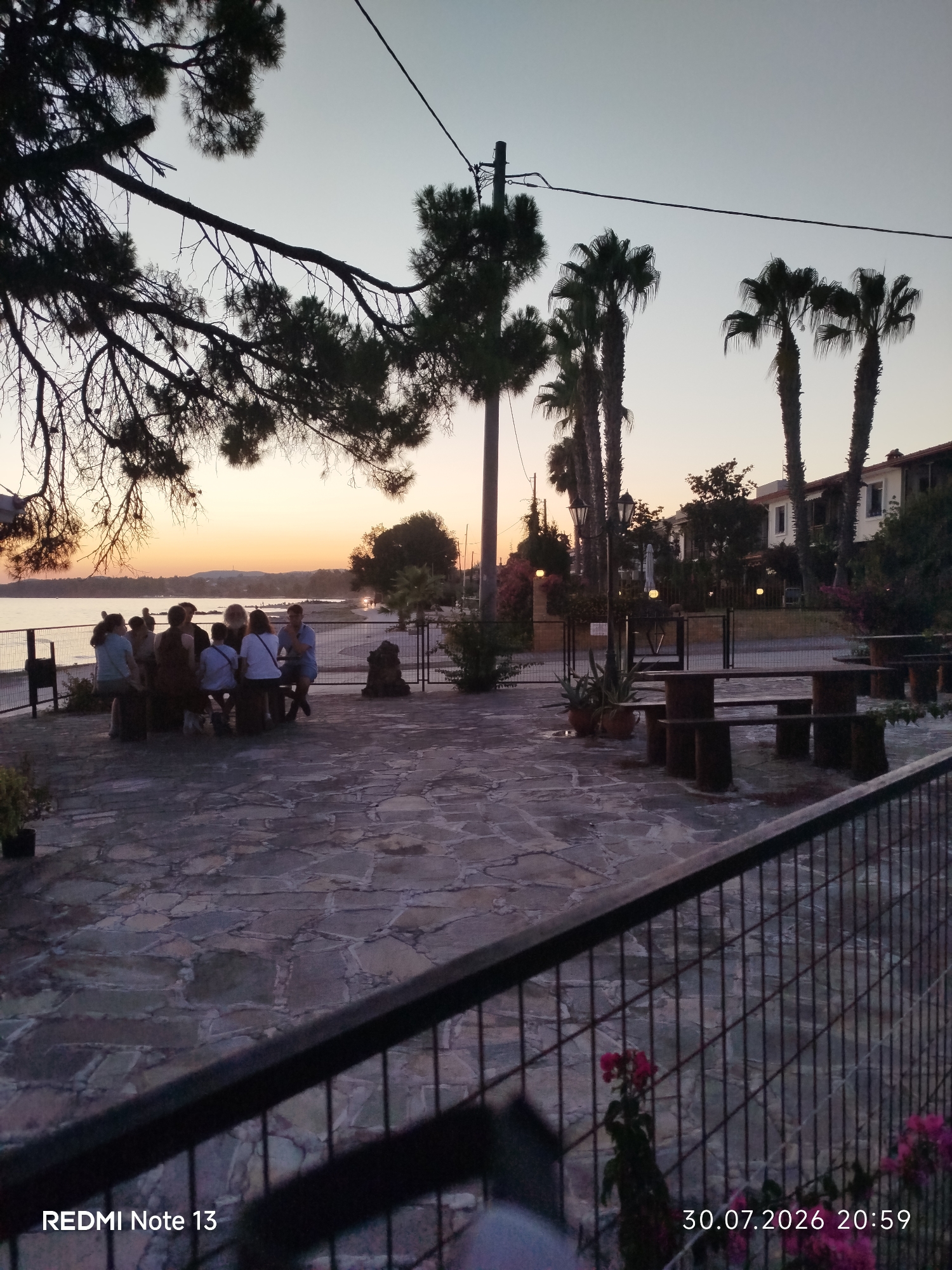 The image depicts an outdoor seating area with several people gathered around a table by a body of water, likely a beach or bay, in Sithonia Municipal Unit, Greece. The time appears to be dusk, with a warm orange glow from the setting sun visible on the horizon. Tall palm trees and other vegetation are present, as are some buildings in the background. The ground is paved with flagstones, and a metal fence runs across the foreground. The overall scene suggests a relaxed evening gathering.