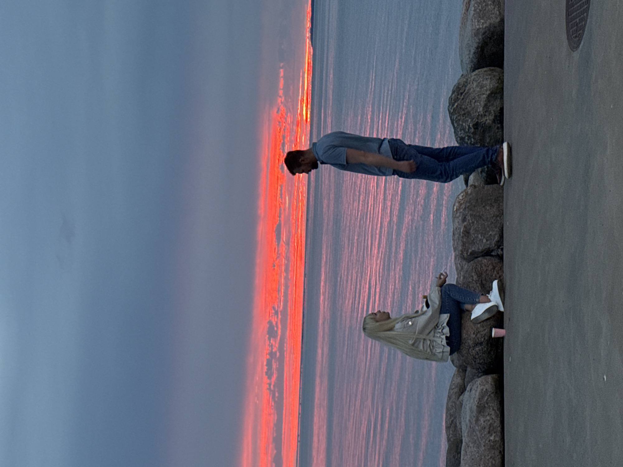 The image captures a serene evening scene at the Tallinn coast in Estonia, as the sun dips below the horizon, painting the sky with vibrant hues of red and orange. In the foreground, two individuals are depicted against the backdrop of a calm sea. A woman with long blonde hair is seated on large rocks, wearing a light-colored jacket and blue jeans. Beside her, a pink cup is placed on the rocky surface. Facing her is a man with short brown hair, dressed in a blue polo shirt and blue jeans, standing a few feet away. The sea stretches out to the horizon, reflecting the fiery colors of the sunset, while the sky above is a gradient of blue and grey. The overall ambiance is tranquil and picturesque, suggesting a moment of quiet contemplation or conversation by the sea.