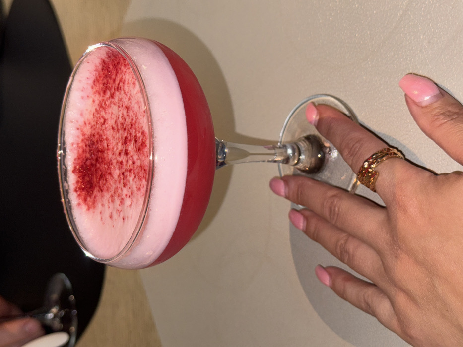 The image shows a vibrant pink and red cocktail in a coupe glass, held by a person's hand. The glass is positioned in the upper center of the frame, held by fingers gripping the stem, with the base resting on a light-colored surface.
The cocktail itself has two distinct layers. The bottom layer is a rich, dark pink or red liquid. Above it, there's a thick, light pink or white foamy layer that fills the top part of the glass. This foamy top is generously garnished with a sprinkling of finely crushed red particles, possibly dried berries or powder, which stand out against the pale foam. Small bubbles are visible on the surface of the foam.
A hand, likely a woman's, is holding the clear stem of the glass. The nails on the visible fingers are manicured and painted with a light pink or nude polish. On the index finger of the hand, a gold-toned ring with an irregular, textured, or organic design is worn.
The glass and hand are resting on a plain, light beige or cream-colored table surface. In the blurred background to the left, a dark, indistinct shape is visible, possibly another table or piece of furniture. Another glass is partially visible in the bottom left corner, suggesting another drink or person nearby. The lighting appears to be indoor and somewhat dim, casting subtle shadows and highlighting the colors of the cocktail.