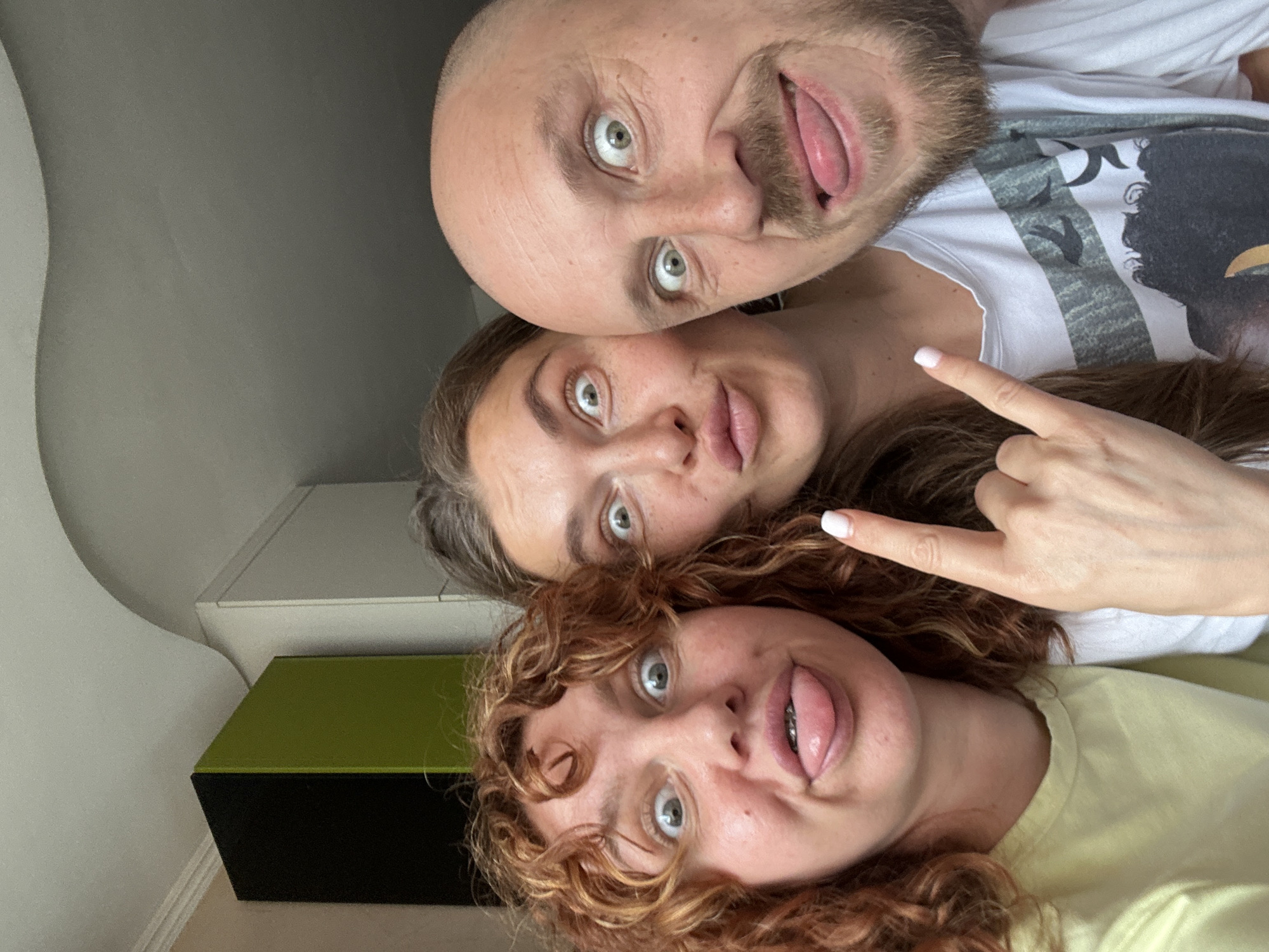 Three individuals are in the frame in an indoor setting. A bald man with a beard is at the top, sticking his tongue out. Below him is a woman with long brown hair making a hand gesture. At the bottom is a woman with curly red hair, also sticking her tongue out. A white cabinet and a green and black object are visible in the background. The location is unknown, and there is no evidence to support that it is in Sochi, Russia.