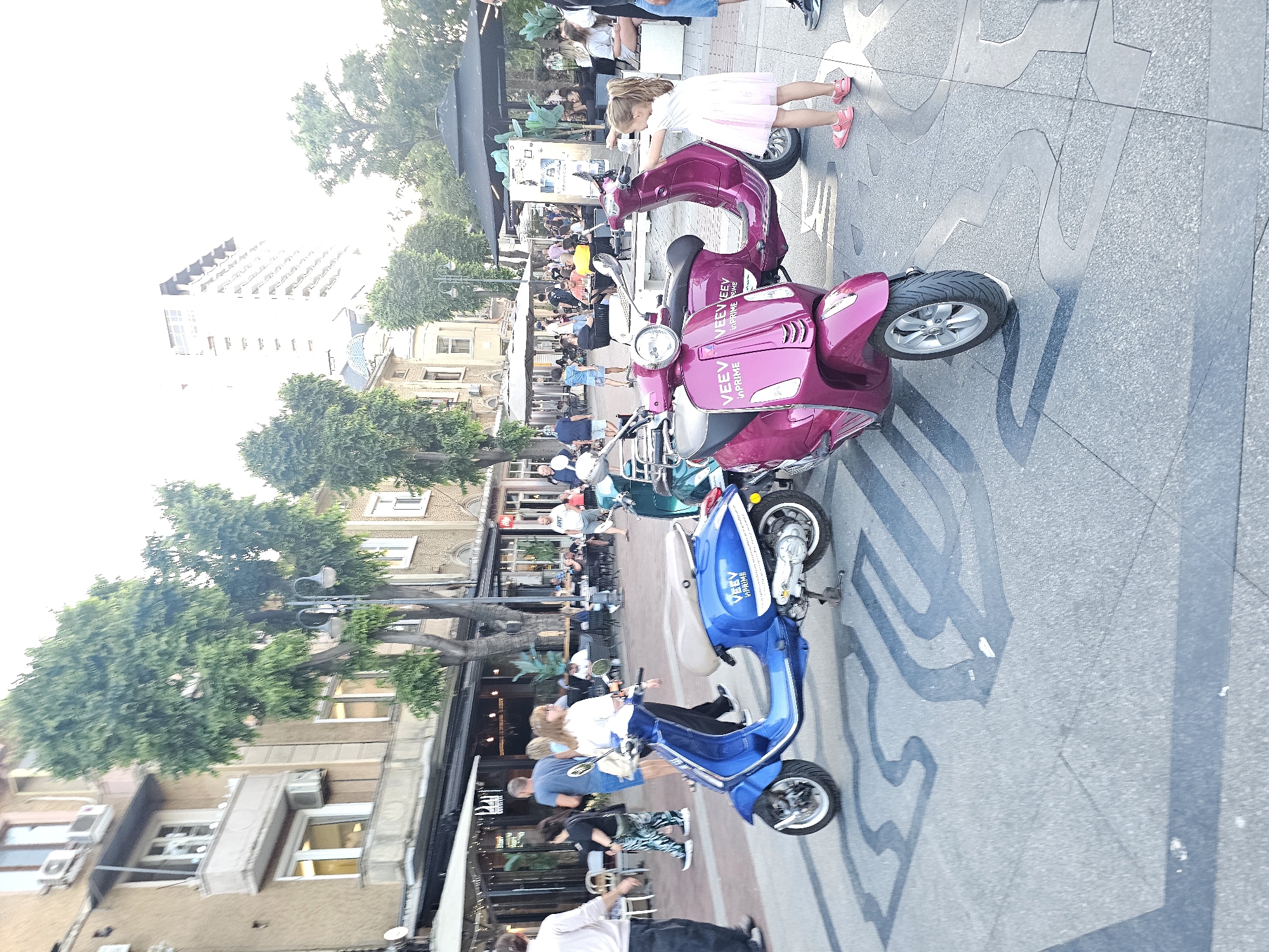 In an outdoor urban setting, two parked electric scooters, one blue and one magenta, are the central focus. The blue scooter is on the left and the magenta scooter is on the right. A young girl in a pink dress and sandals stands to the right of the magenta scooter, with her right arm extended as if interacting with it. In the background, people are visible walking and seated at outdoor tables in front of buildings that appear to be shops or restaurants. The ground is paved with square tiles and features a decorative inlay. Trees are present throughout the scene, and a tall, modern building is visible in the distance. The lighting suggests it is daytime, with a bright sky.