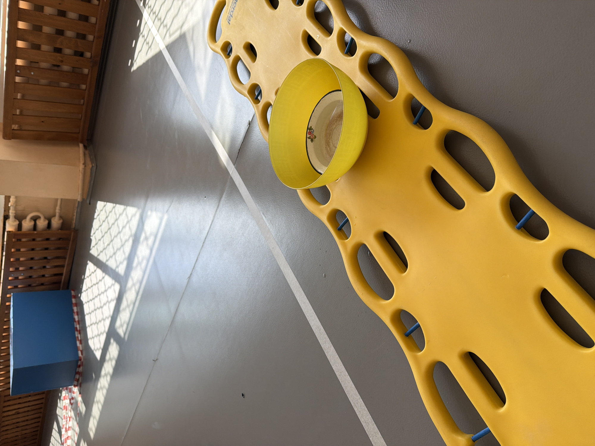 The image captures an indoor scene, likely a gymnasium or institutional hallway, under bright daylight. A prominent yellow plastic object, resembling an emergency spine board or a large bodyboard, lies diagonally across a light gray floor. This board features numerous oval-shaped cutouts that serve as handholds, and blue fasteners or straps are visible through some of them. Partially visible text on the board suggests a brand name, possibly beginning with "Aerda" or "Sterdal".
Resting on the yellow board is a bright yellow plastic bowl, placed upside down. Beneath it, a smaller, white ceramic or enamel bowl is visible, featuring a subtle floral pattern with red and green elements on its base.
The floor is a uniform light gray, traversed by a crisp white stripe, indicating a marked area or pathway. Strong sunlight streams in from the left, casting distinct grid-like shadows onto the floor and a gray wall, suggesting light filtering through a window with a grid pattern or a mesh fence.
To the far left, a section of a wooden slatted structure, possibly shelving or a decorative screen, is visible. Below it, a beige structure and several white cylindrical objects are partially seen. Further left, a blue rectangular box-like object, possibly a piece of equipment or furniture, stands next to the wooden structure, with a red and white checkered pattern (possibly fabric or tape) draped on one side. No human figures are present, and no specific activity is taking place; the scene appears static.
