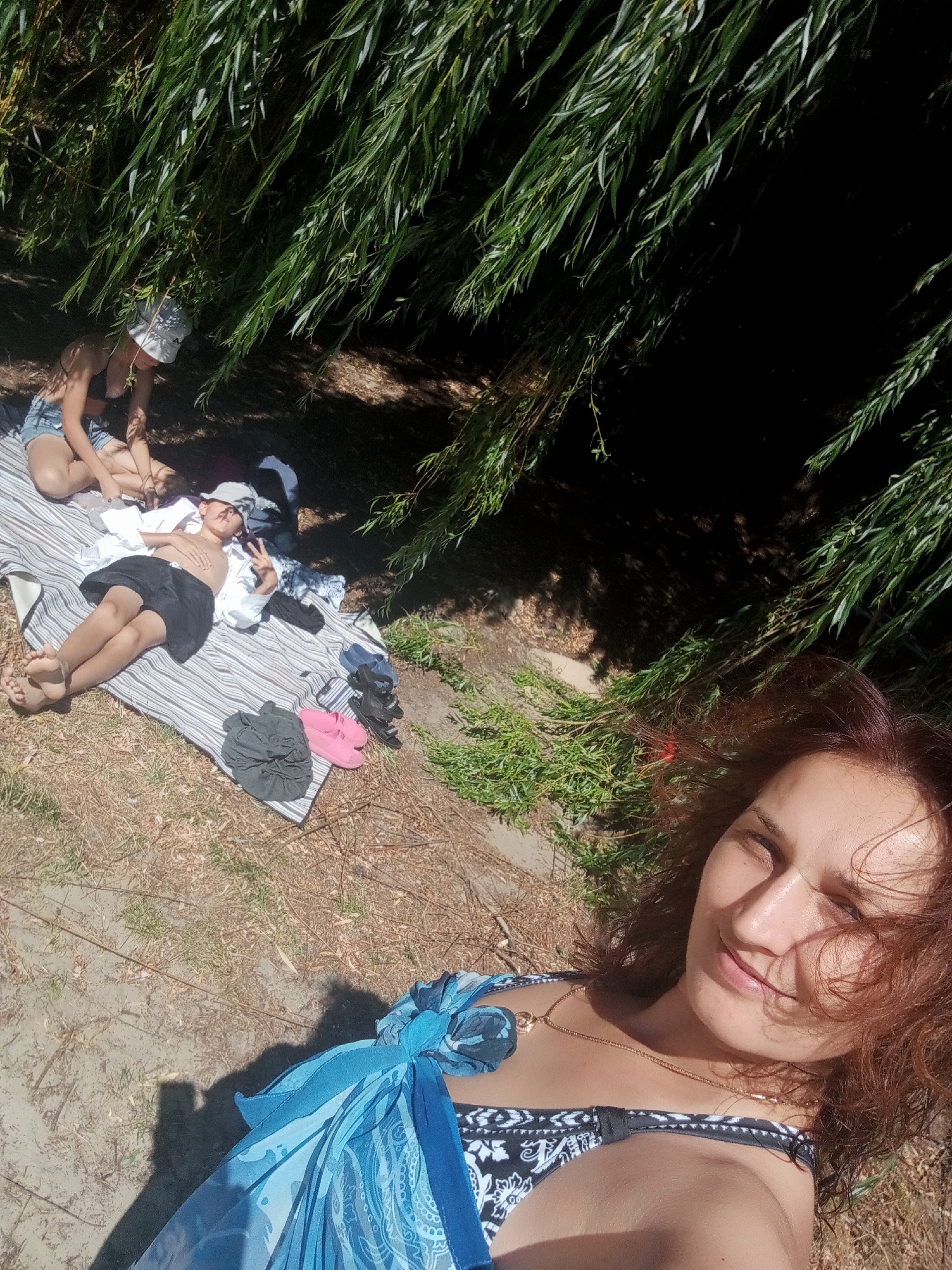 A woman lies on the ground, her face close to the camera and partially obscured by her auburn hair. She wears a black and white patterned bikini top and a blue sarong. Behind her, two people are picnicking under the shade of willow trees. One person, wearing denim shorts and a white hat, sits on a striped blanket, while another, a child wearing a white shirt and black shorts, lies on their back on the same blanket, giving a peace sign with their fingers. Several pairs of sandals and folded clothing are scattered on the ground near the blanket. The scene is set in a park or natural area, possibly near a body of water, given the sandy ground and lush vegetation. The lighting suggests it is a sunny day. There is no specific indication of the city of Obariv, Ukraine, in the image.