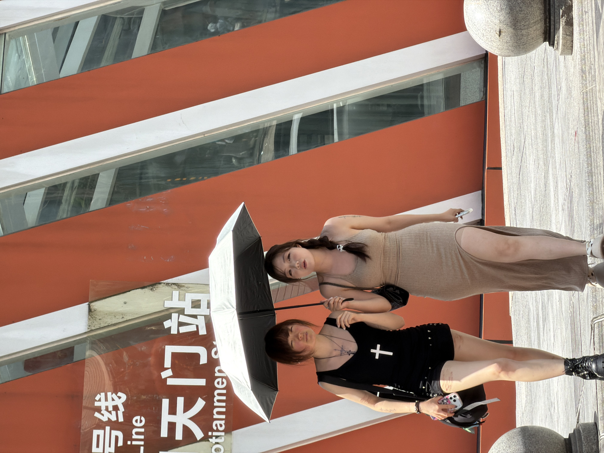 Two young women walk under a black umbrella on a sunny day in the Yuzhong District of China. The woman on the left wears a black sleeveless top with a large white cross pendant and black shorts, while the woman on the right wears a form-fitting beige dress with a high slit. Both women are carrying purses. Behind them, a modern building features bright orange and white diagonal stripes and large windows reflecting the surroundings. The ground is paved with grey tiles, and a large, spherical stone monument is visible on the right side of the frame. The scene appears to be a street or public walkway in a city.