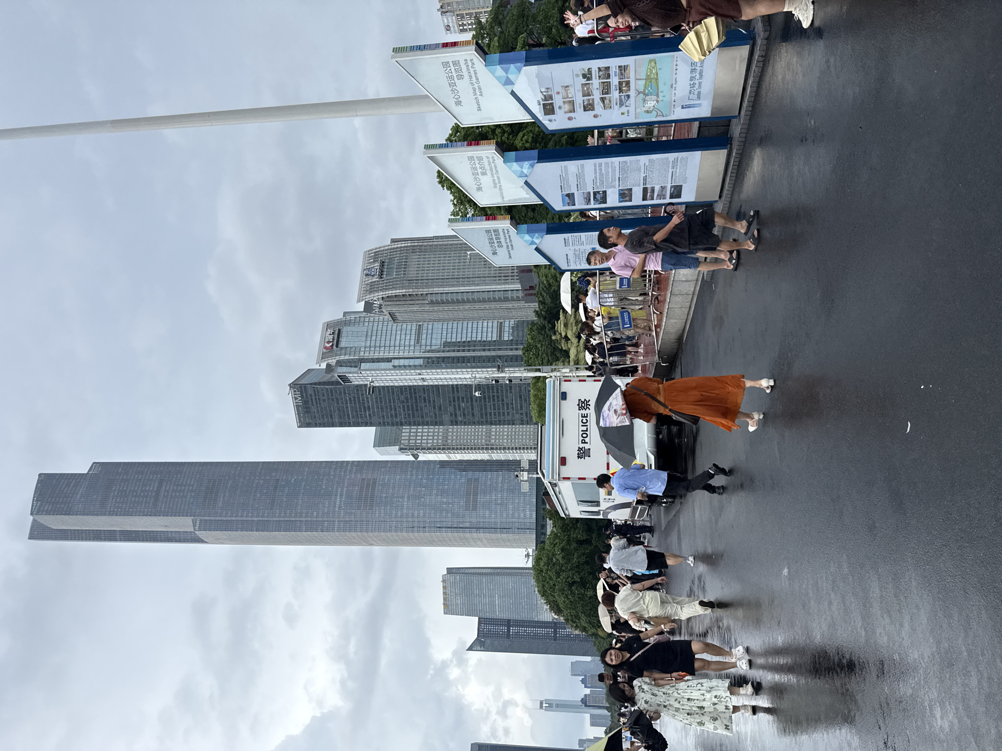 In Tianhe District, China, on a wet paved surface, a group of people are walking under a cloudy sky. In the background, several modern skyscrapers, including a very tall, slender, dark grey one with a prominent vertical division, dominate the skyline. To the right of the frame, a tall, white pole extends upwards. In the middle ground, a white police van with "POLICE" written in both English and Chinese characters is parked. Several informational display boards with blue borders are also present, featuring text and images, one of which is titled "Sketch Map of Haixinsha Asian Games Park." People are scattered throughout the scene, some holding umbrellas, suggesting it is raining or has recently rained. The wet ground reflects the surrounding buildings and the overcast sky.