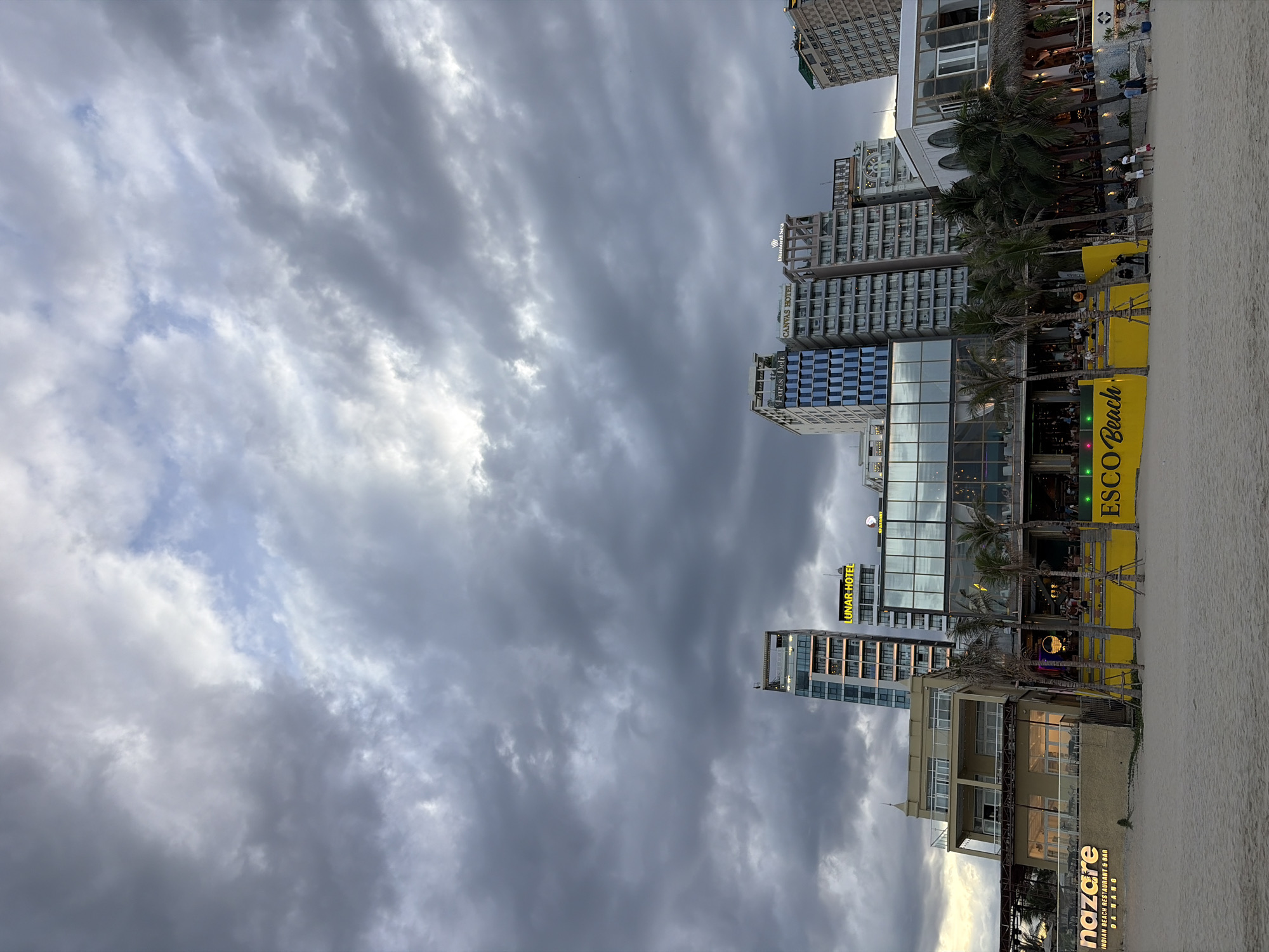 The image depicts a beachfront scene in Da Nang, Vietnam, under a dramatic, overcast sky. In the foreground, a sandy beach stretches across the frame. Behind the beach, a strip of buildings and establishments line the shore.
Several multi-story buildings rise into the sky, some with modern glass facades and others with more traditional architectural elements. Signs on these buildings indicate hotel names such as "LUNAR HOTEL," "Paris Deli," and "CANVAS HOTEL." Below the taller buildings, lower-rise structures house restaurants and bars, identifiable by their illuminated signs and outdoor seating areas. One sign prominently displays "NAZARE INDIAN BEACH RESTAURANT & BAR DA NANG." Another large yellow sign reads "ESCO Beach." Palm trees are interspersed among the buildings, adding to the tropical atmosphere.
People are visible on the beach and in front of the establishments, appearing to be patrons or staff. Some individuals are walking on the sand, while others are gathered near the entrances of the businesses. The overall scene suggests a lively area, likely a tourist destination, despite the cloudy weather. The lighting is subdued, indicating it may be late afternoon or early evening, or a day with significant cloud cover.