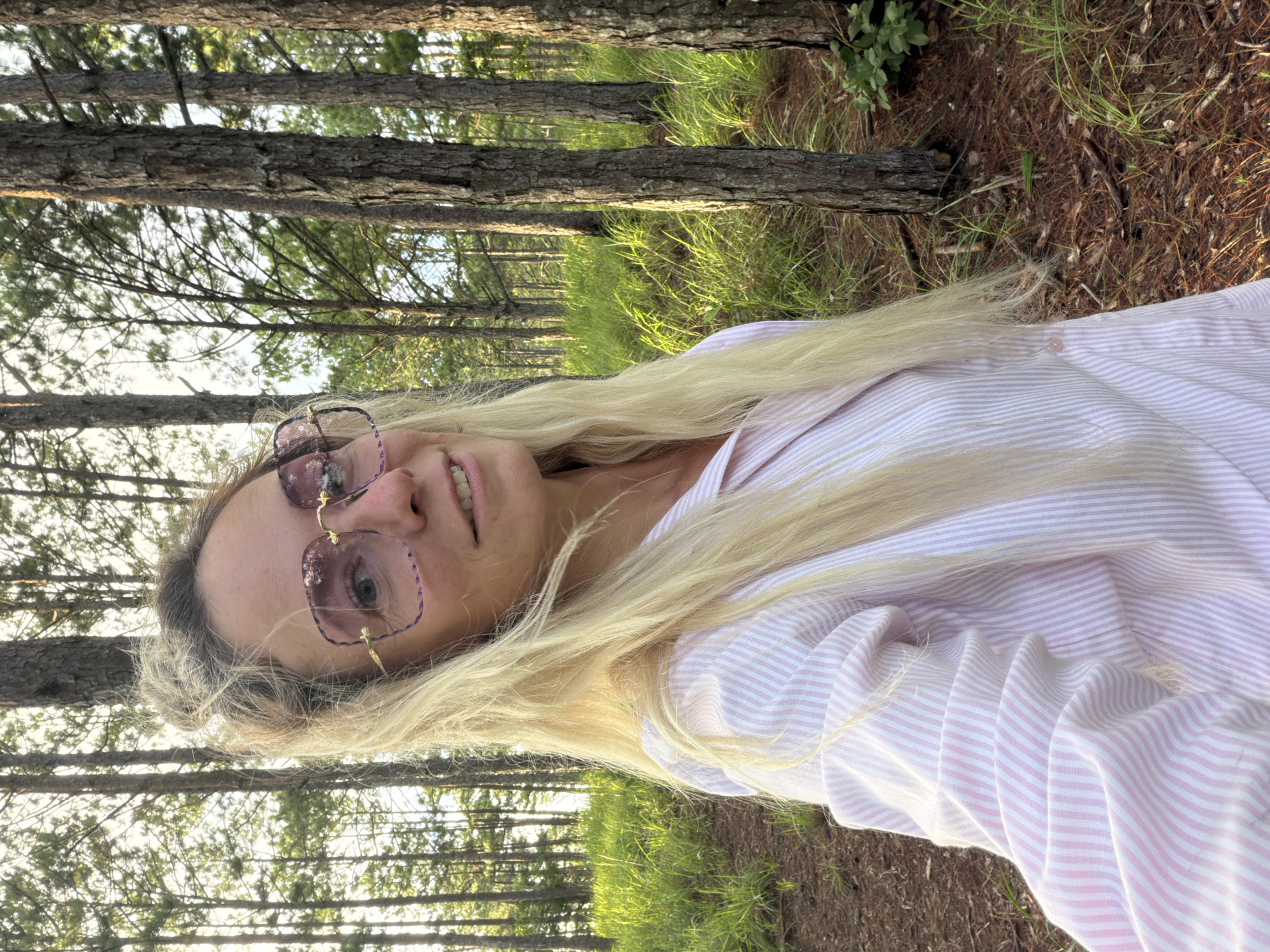 A blonde woman wearing large pink-tinted glasses and a pink and white striped button-down shirt is taking a selfie in a forest. Her hair is long and appears windswept. The background shows several tall, slender trees with rough bark, and lush green grass and pine needles on the ground. The lighting suggests it is daytime, possibly late afternoon due to the warm glow. The location is the city of Xuan Truong - Da Lat, Vietnam.