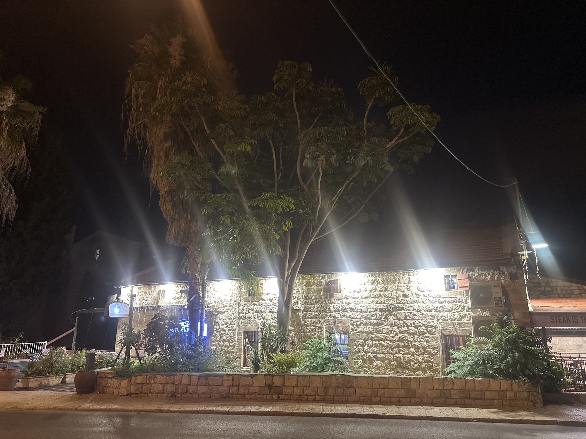 This nighttime photograph captures a rustic, stone building illuminated by artificial lights, suggesting it is likely a restaurant or establishment open after dark. The building has a traditional stone facade with visible windows and air conditioning units. A prominent tree with dense foliage stands in front of the building, its branches spread wide. To the left, a tall palm tree adds to the tropical or Mediterranean feel of the scene.
The foreground shows a paved road and a low stone wall bordering a planter area filled with greenery. The lighting is dramatic, with bright spotlights creating strong highlights and lens flares, indicating a clear night. A sign hanging from the building reads "STEAK IS TH". Another sign visible further left has the text "Galilee Grill" and "Savor".
The overall atmosphere is inviting, with the warm glow of the lights contrasting with the dark night sky. The scene is set in Haifa, Israel, a city known for its coastal beauty and diverse architecture. The time of day is clearly night, and there are no indications of specific weather conditions other than the darkness. No people are visible in the frame, making the focus solely on the architecture, vegetation, and the inviting ambiance of the establishment.