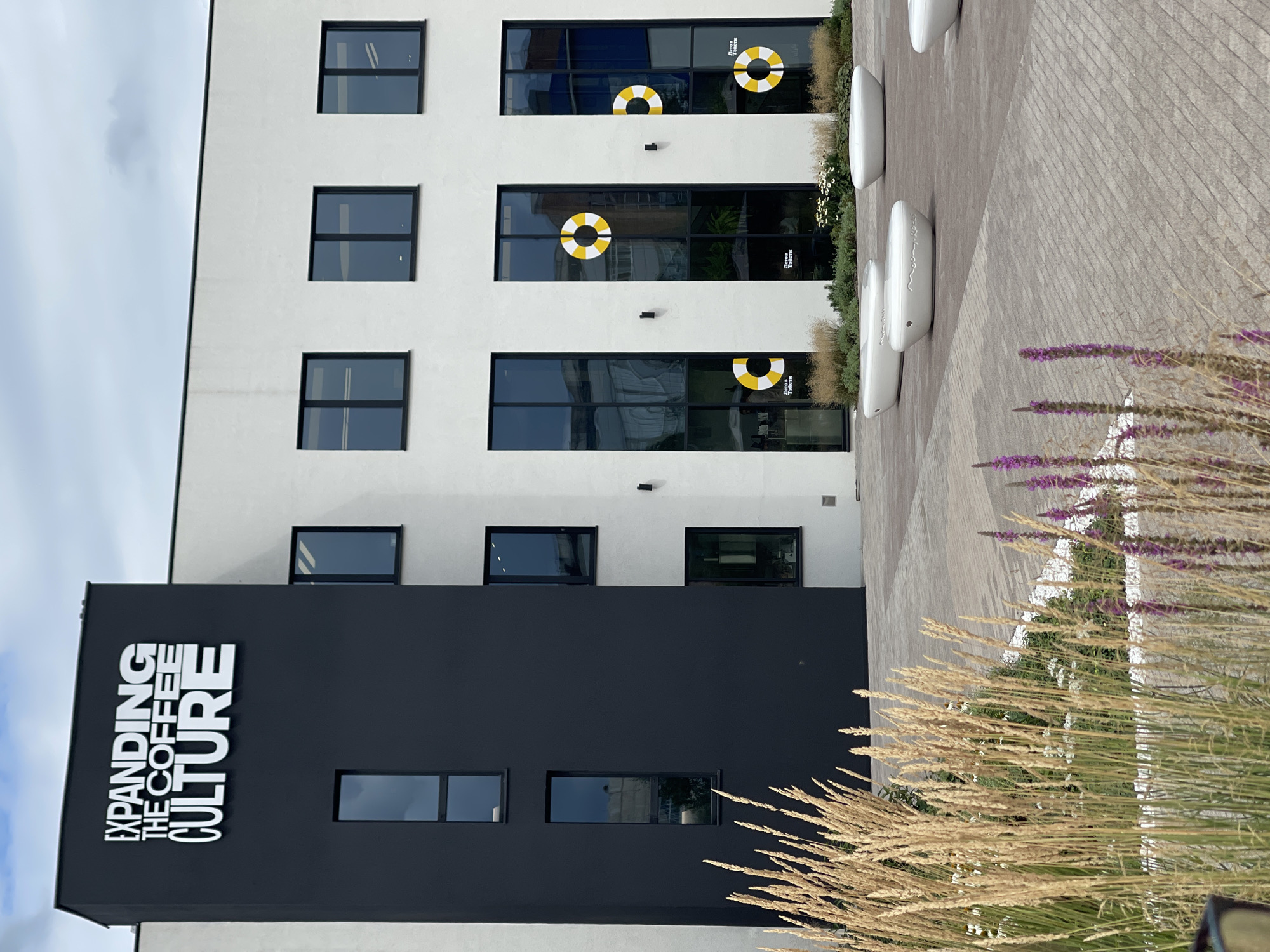 The image shows the exterior of a modern building with white and dark grey facades. The building has several large windows, some of which have yellow and white circular decals resembling life preservers. The text "EXPANDING THE COFFEE CULTURE" is prominently displayed in white lettering on the dark grey section of the building. In the foreground, there are planters with dry grasses and some purple flowers. Several white, curved benches are placed on the paved area in front of the building. The sky is cloudy. This building is located at the intersection of Komsomolsky Prospekt and Gagarina Street in Izhevsk, Russia, and is part of the "Coffee House" development.