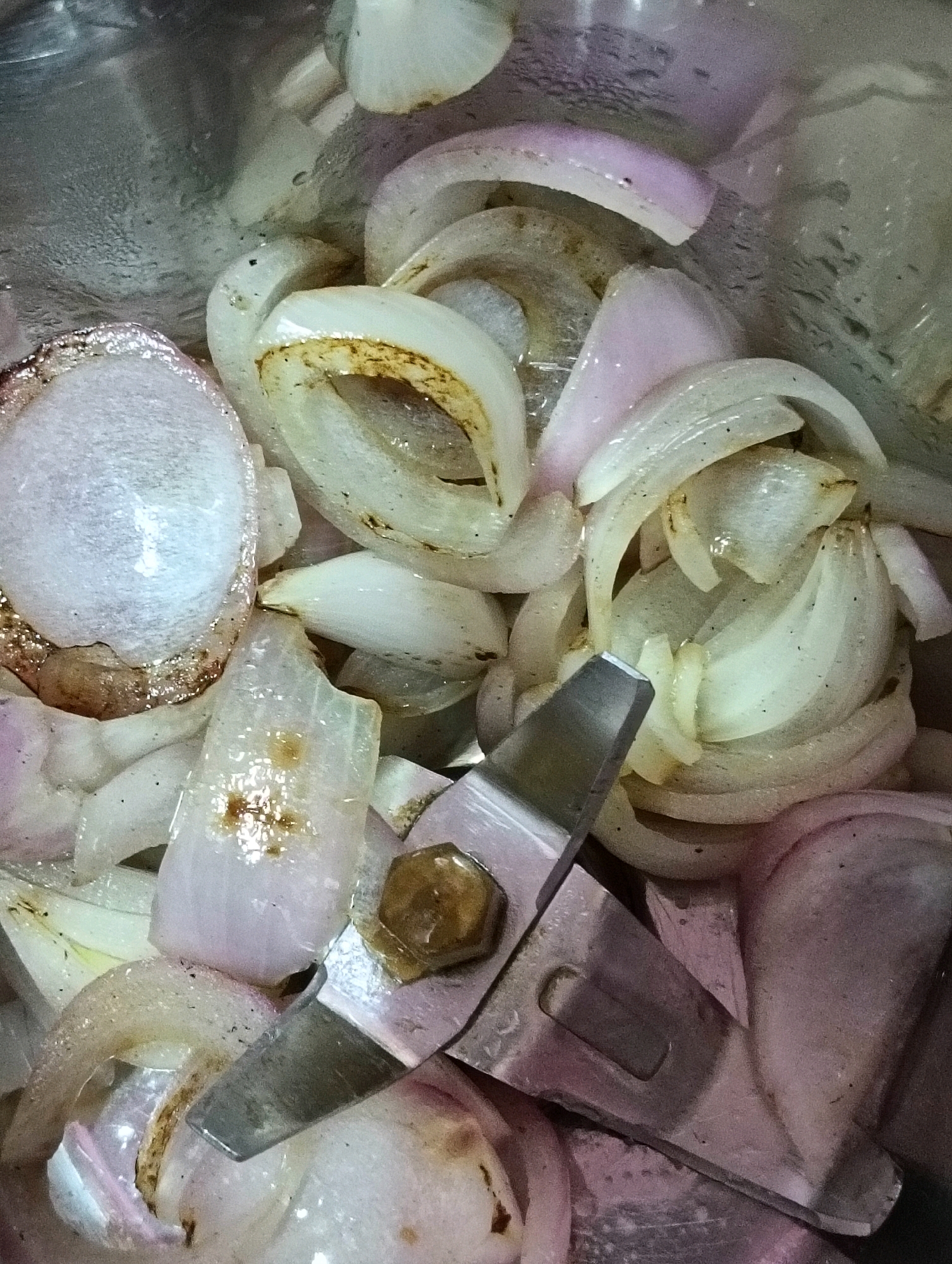 The image displays chopped red onions inside a blender jar, with the blender's blade visible in the foreground. The onions are cut into irregular pieces and slices, some with visible purple skin and others primarily white. The blender jar appears to be made of stainless steel, with some condensation on its inner surface. The scene is tightly cropped, focusing on the contents of the blender. There are no people or animals visible in the image. The overall impression is a close-up view of food preparation ingredients within a kitchen appliance. The location of Hosapete, India is not discernible from the image itself.