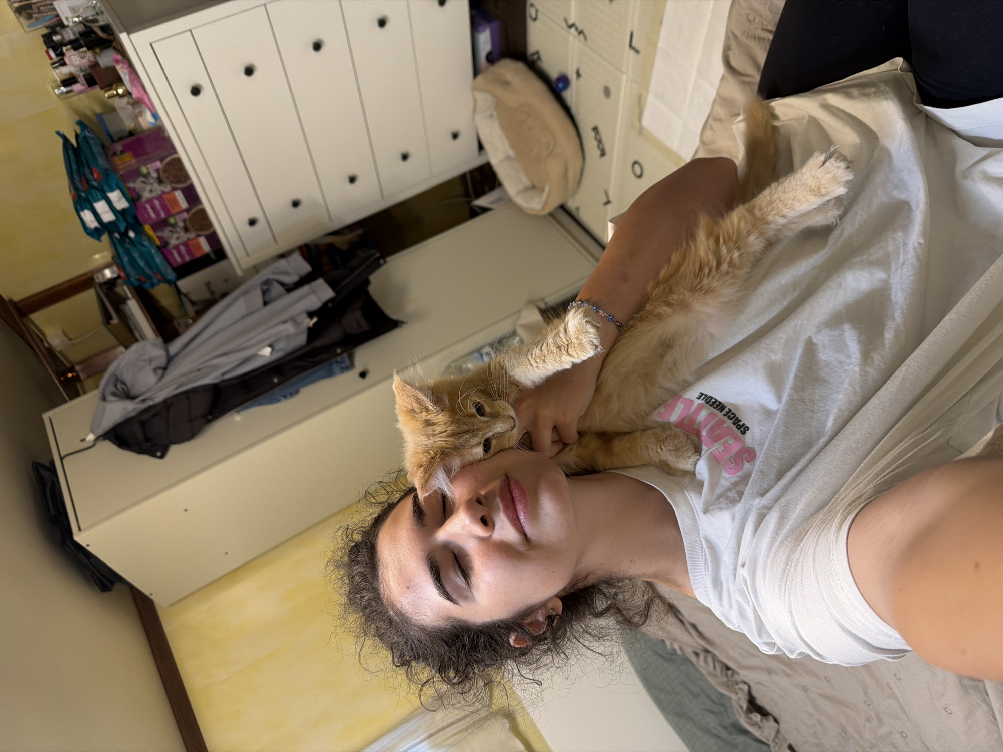 A young woman with curly brown hair and closed eyes is holding an orange kitten. She is wearing a white t-shirt with pink lettering that reads "SEATTLE SPACE NEEDLE." The kitten is nestled against her neck and shoulder. They are indoors, in what appears to be a bedroom. In the background, there is a white dresser with drawers, a closet with hanging clothes, and a pet bed on the floor. The walls are a pale yellow. The scene does not provide any specific indicators of location in the city of Sofia, Bulgaria.