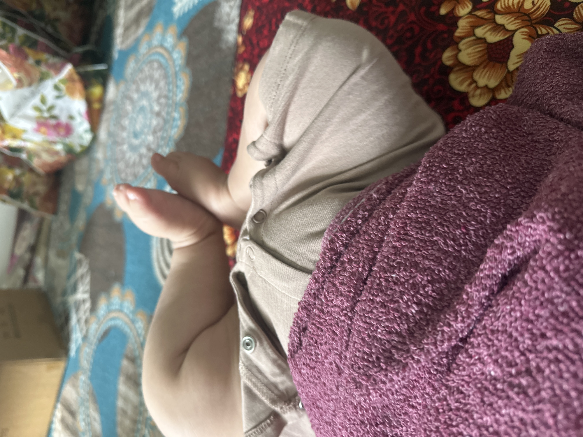 The image is a close-up shot of a baby's legs and torso, partially covered by a purple towel. The baby is wearing a light brown onesie with snaps. The background shows a colorful patterned blanket or rug, with floral motifs and circular designs in blue and grey. To the left, a portion of another patterned fabric, possibly a bag or cushion with pink and yellow flowers, is visible. The scene appears to be indoors, likely on a bed or couch, given the textiles. The lighting suggests daytime. The overall impression is intimate and domestic, focusing on the soft textures and the delicate details of a baby's body. There is no visible text in the image. The location context provided is Тайгараев, Kyrgyzstan.