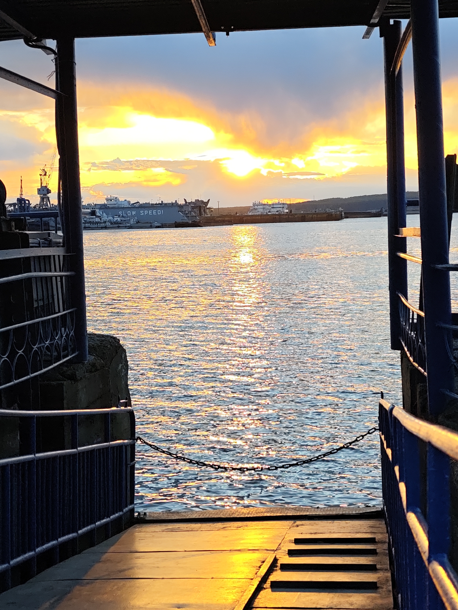 The image shows a sunset over the Kama River in Chaikovsky, Russia, as viewed from a dock or landing. The sky is filled with yellow and orange hues as the sun dips below the horizon, reflecting on the water's surface. Several vessels, including a large barge labeled "SLOW SPEED!" and cranes on the shore, are visible in the background. The foreground includes the dock's railing, posts, and steps leading down to the water, casting shadows due to the low sunlight.