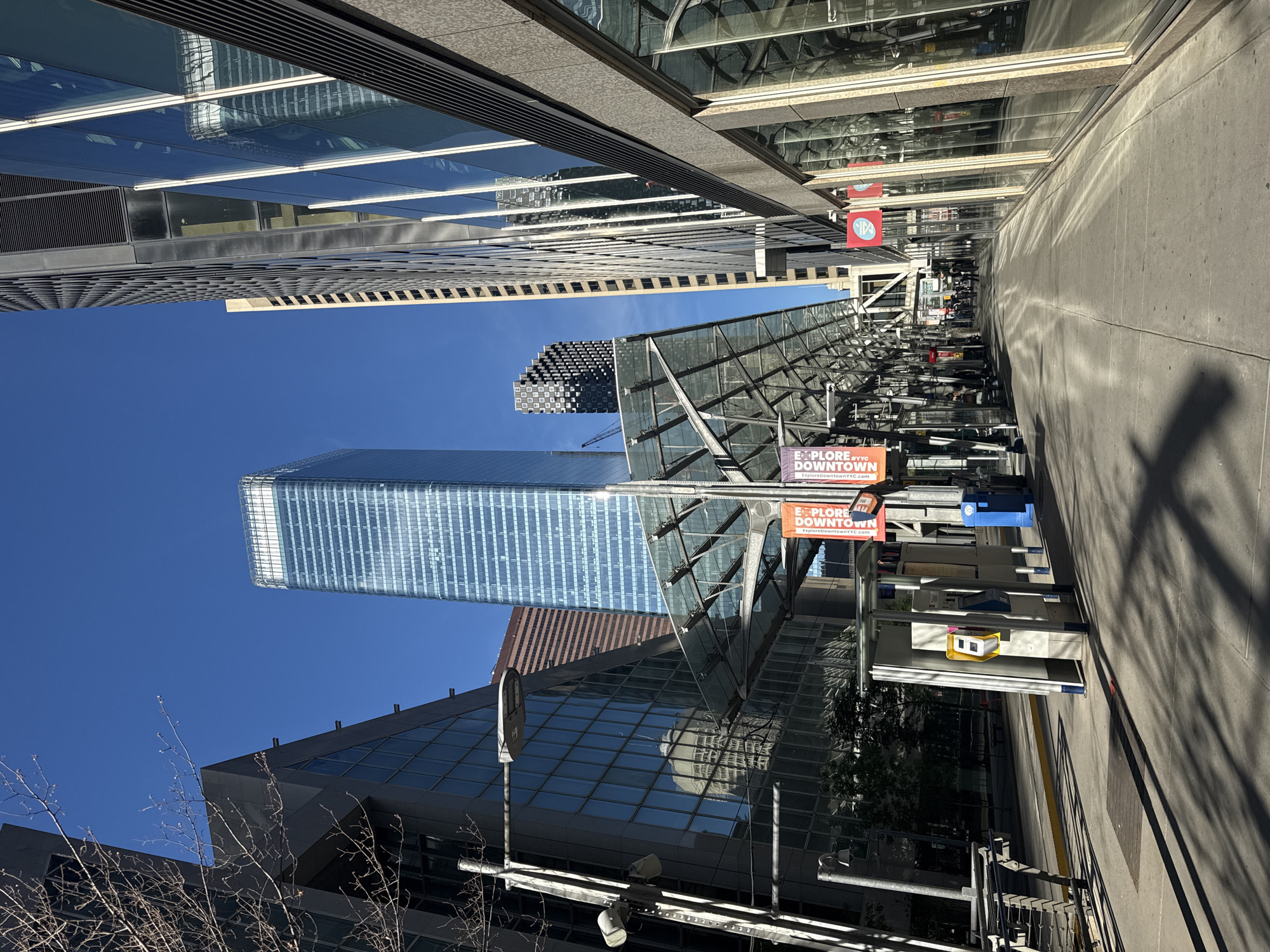 The image shows a sunny day in downtown Calgary, Canada, with a clear blue sky. The scene is dominated by modern skyscrapers with glass facades, reflecting the surrounding buildings and the sky. In the foreground, there is a pedestrian walkway lined with buildings and a transit station with a glass canopy. The station features shelters, ticket machines, and trash receptacles. Banners advertising "EXPLORE DOWNTOWN" are visible, suggesting a vibrant urban area. The shadows cast by the buildings and the canopy indicate the time of day. The overall impression is one of a bustling, contemporary city.