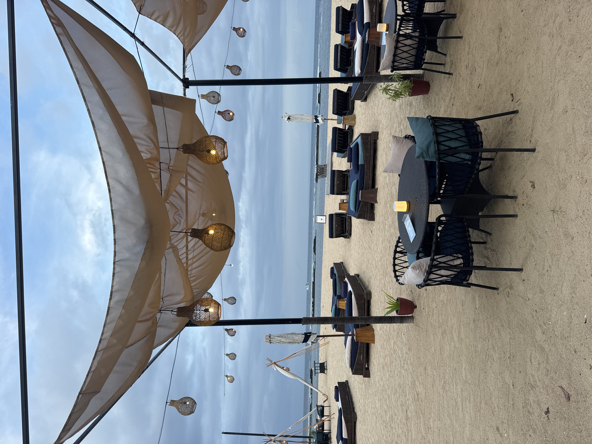 This image depicts a beach scene in Kuta Selatan, Indonesia. The foreground consists of a sandy beach with patio furniture, including tables and chairs made of dark wicker material. Pillows provide comfort on the seating. Planters with greenery are situated near the furniture sets. Overhead, a large awning or sunshade is suspended by dark metal poles, and decorative hanging lights illuminate the area. The backdrop is the ocean and sky, showing a cloudy, overcast day. In the midground, beach loungers with blue cushions are lined up along the shoreline. An assortment of white umbrellas are sparsely erected among the loungers. The horizon indicates calm water. The overall scene suggests a relaxed, beachfront resort or dining area.