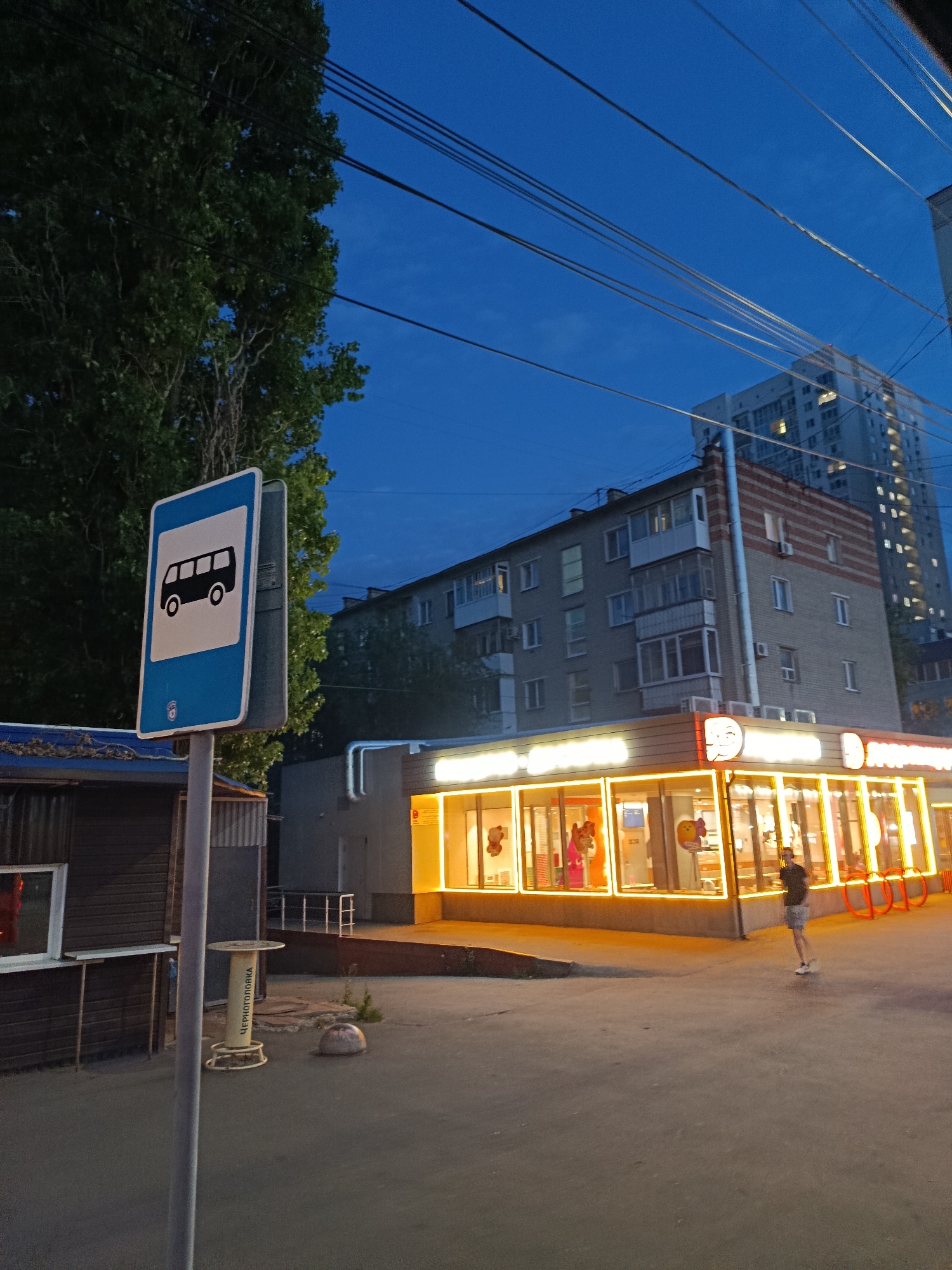 The image depicts a bus stop sign in the foreground, with a stylized bus symbol on a blue and white background. Behind the sign, a dark-colored, one-story building with a corrugated metal facade is visible on the left. To the right of the bus stop, a brightly lit, modern establishment with large glass windows and illuminated signage, possibly a fast-food restaurant or cafe, is the central feature of the middle ground. A single person, wearing a dark t-shirt and light-colored shorts, is walking away from the camera in front of this establishment. In the background, two multi-story apartment buildings are visible against a twilight blue sky. Overhead, power lines stretch across the sky. The scene is likely a street in the city of Saratov, Russia, given the presence of a bus stop sign with Cyrillic text on a nearby spool of wire. The overall atmosphere is that of an urban environment transitioning from day to night, with artificial lights illuminating the street.