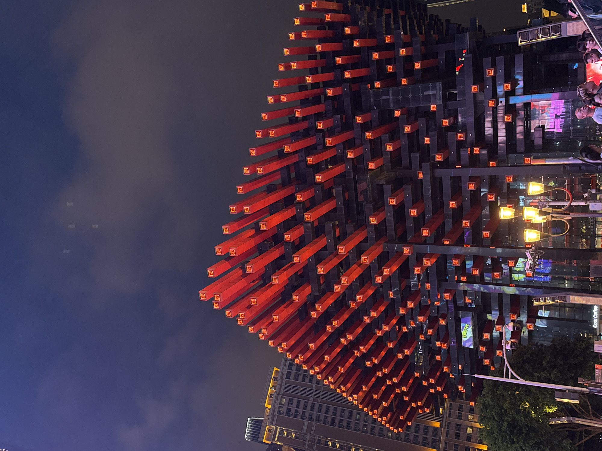 The image shows the exterior of the Hongyadong complex at night in Chongqing, China. The structure is characterized by a prominent, multi-tiered facade made of dark materials interspersed with numerous red, rectangular light fixtures that emit a warm glow. These lights are arranged in a grid-like pattern across the building's exterior. The sky above is dark and cloudy, with faint hints of light pollution. To the left of the main structure, a tall, modern building with illuminated windows is visible. In the foreground, there are streetlights and a glimpse of trees. A crowd of people are gathered at the base of the complex, some are looking up at the building, and others are walking by. The overall scene suggests a bustling urban environment at night. The Yuzhong District of Chongqing is known for its dramatic topography and unique architecture, and Hongyadong is a popular tourist destination that embodies this.