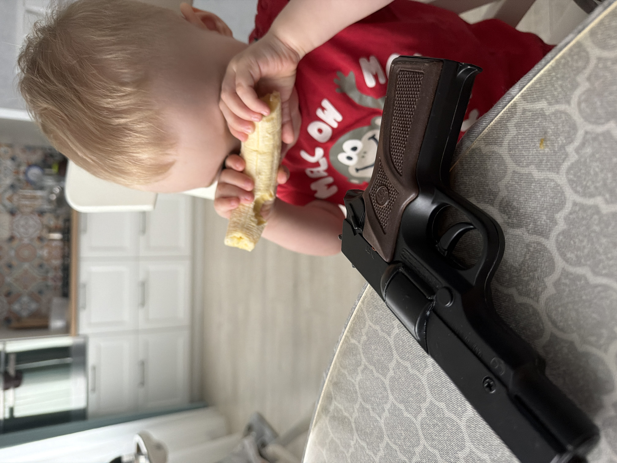 A child with blond hair is eating a banana while sitting at a table. A toy gun is also on the table. The location cannot be determined from the image.