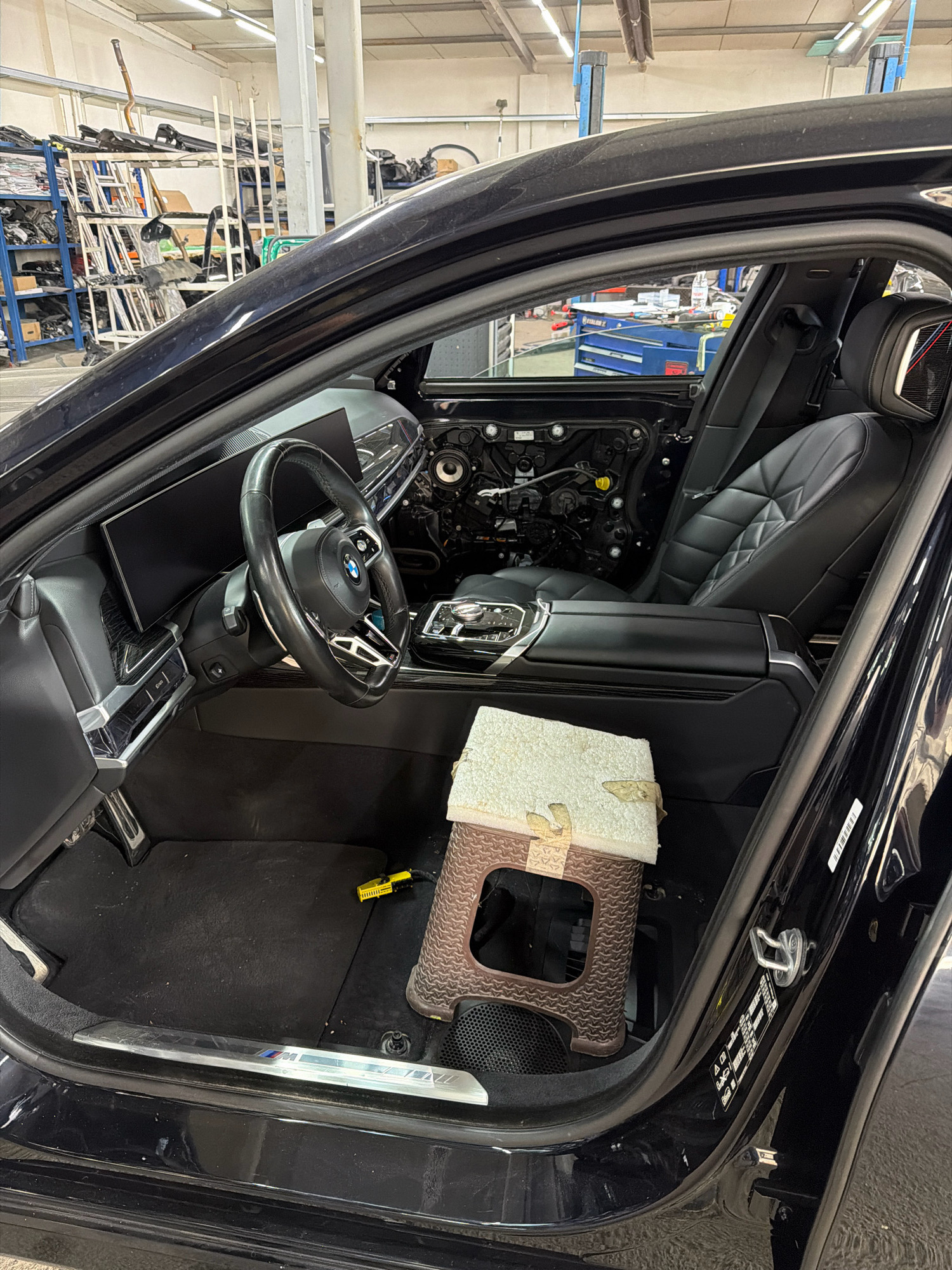 The image depicts the interior of a black car, likely a luxury sedan, undergoing some form of maintenance or repair. The driver's side door is open, revealing the interior. The dashboard features a large, wide digital display, and the steering wheel is visible. The door panel on the driver's side appears to be partially removed, exposing internal components. The car's seats are upholstered in black leather with a quilted pattern. A small, brown plastic stool with a white padded top is placed on the floor in front of the passenger seat. The car is situated in what appears to be a garage or workshop environment, with shelves stocked with automotive parts visible in the background. The lighting suggests an indoor setting with overhead fluorescent lights. The specific location of Burgas, Bulgaria, is not ascertainable from the visual information provided in the image. No individuals are present in the frame.
