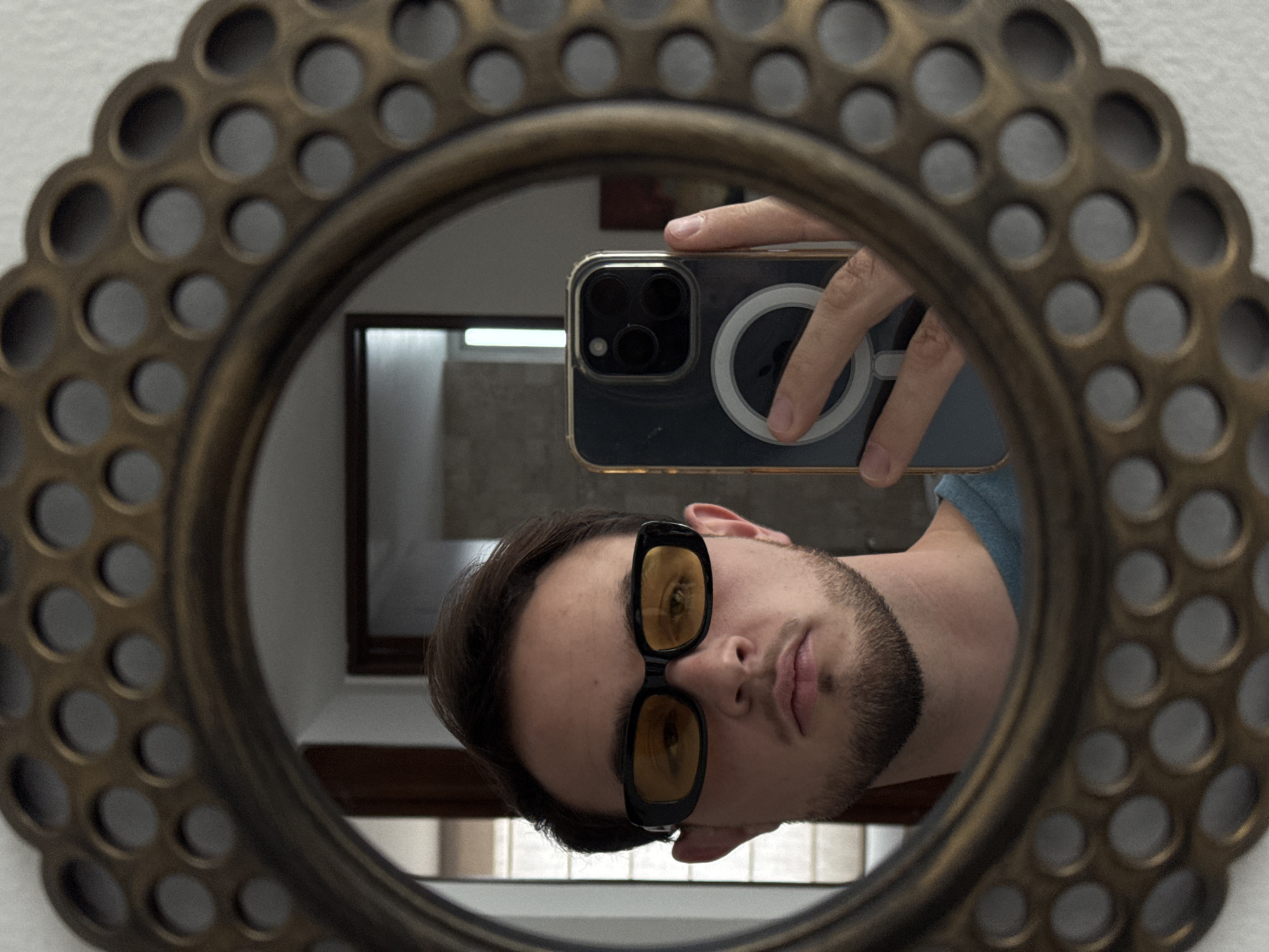A selfie of a man is reflected in a round, ornate mirror with a circular pattern around its outer edge. The man is wearing black-framed sunglasses with yellow lenses. He is holding a smartphone in his hand, which is visible in the reflection. The background includes a window with white blinds. The image was taken in San Bernardino Tlaxcalancingo, Mexico.