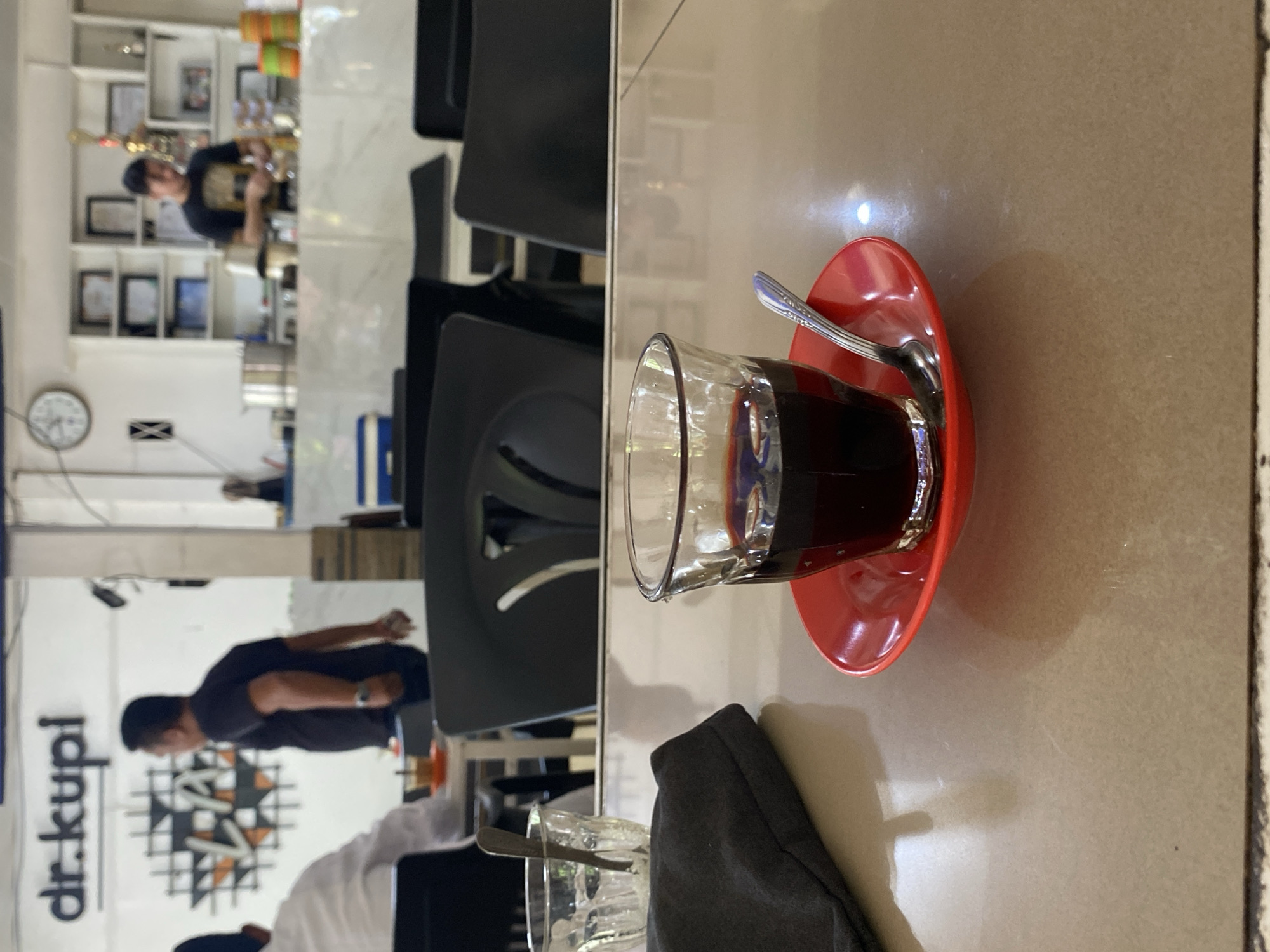 A close-up, eye-level shot shows a small glass of dark coffee on a red saucer with a spoon on a light brown table. The coffee appears to be a local Indonesian brew, likely from Lhokseumawe, Indonesia. In the blurred background, a sign reads "dr. kupi," indicating the establishment is a coffee shop. Behind the counter, a barista is visible, and shelves are stocked with various items. Several patrons are seated at tables, one of whom is facing away from the camera. The overall setting suggests a casual, everyday coffee shop environment.