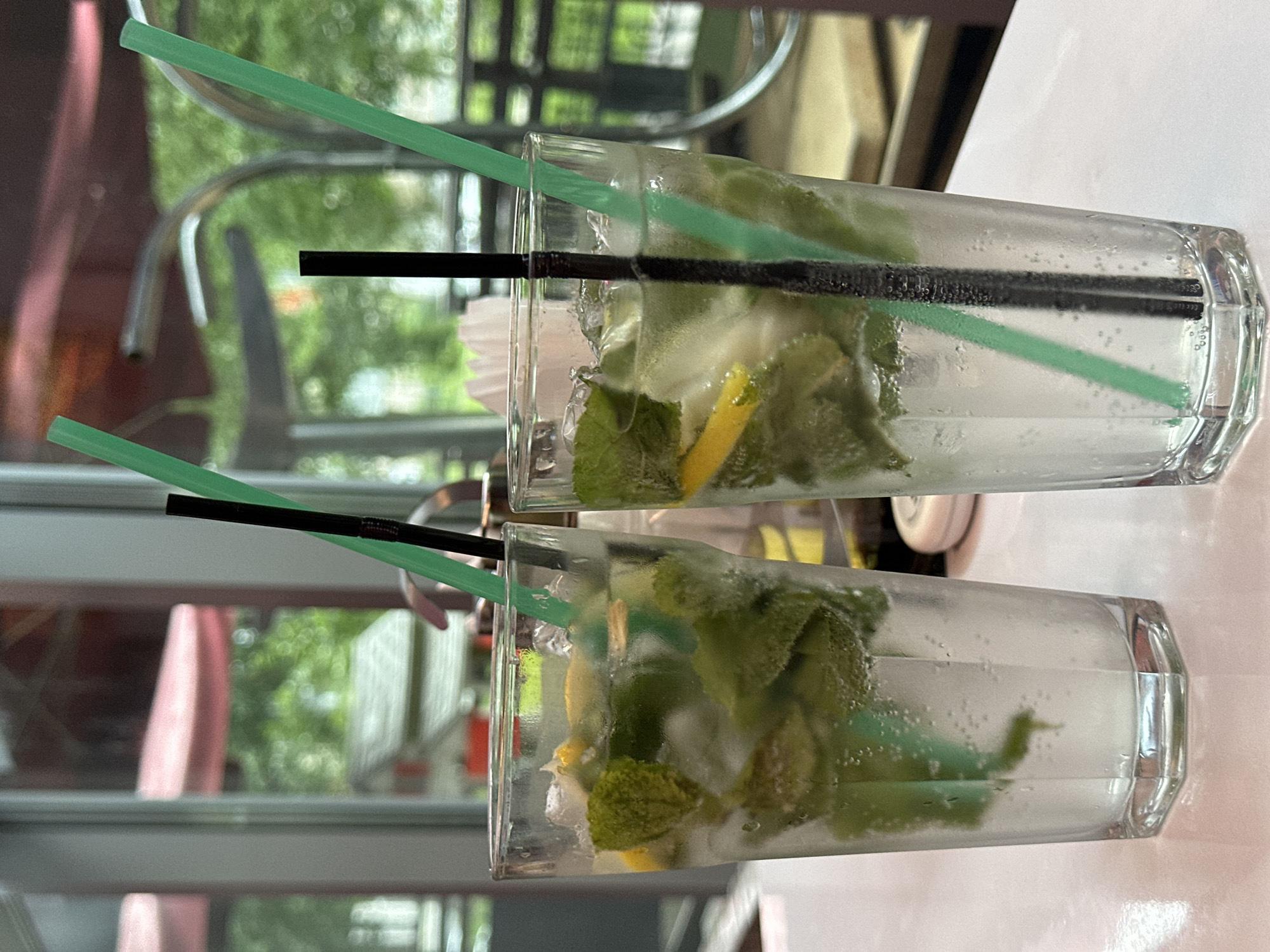 Two glasses filled with a clear, bubbly beverage are the main subjects in this photograph. Each glass contains ice, fresh mint leaves, and wedges of lemon, suggesting a refreshing drink like a mojito. Two straws, one green and one black, are placed in each glass. The glasses are positioned horizontally and appear to be resting on a white surface, possibly a table.
In the background, out-of-focus elements suggest an outdoor or semi-outdoor setting. There are reflections of greenery, trees, and some architectural features like chairs and possibly windows or a building. The lighting indicates it's daytime, with soft light casting shadows. The overall atmosphere is casual and relaxed, hinting at a moment of enjoyment or a break.
There is no visible text in the image. The location context of Chaikovsky, Russia, doesn't offer specific visual cues within the image itself to confirm or deny the location.