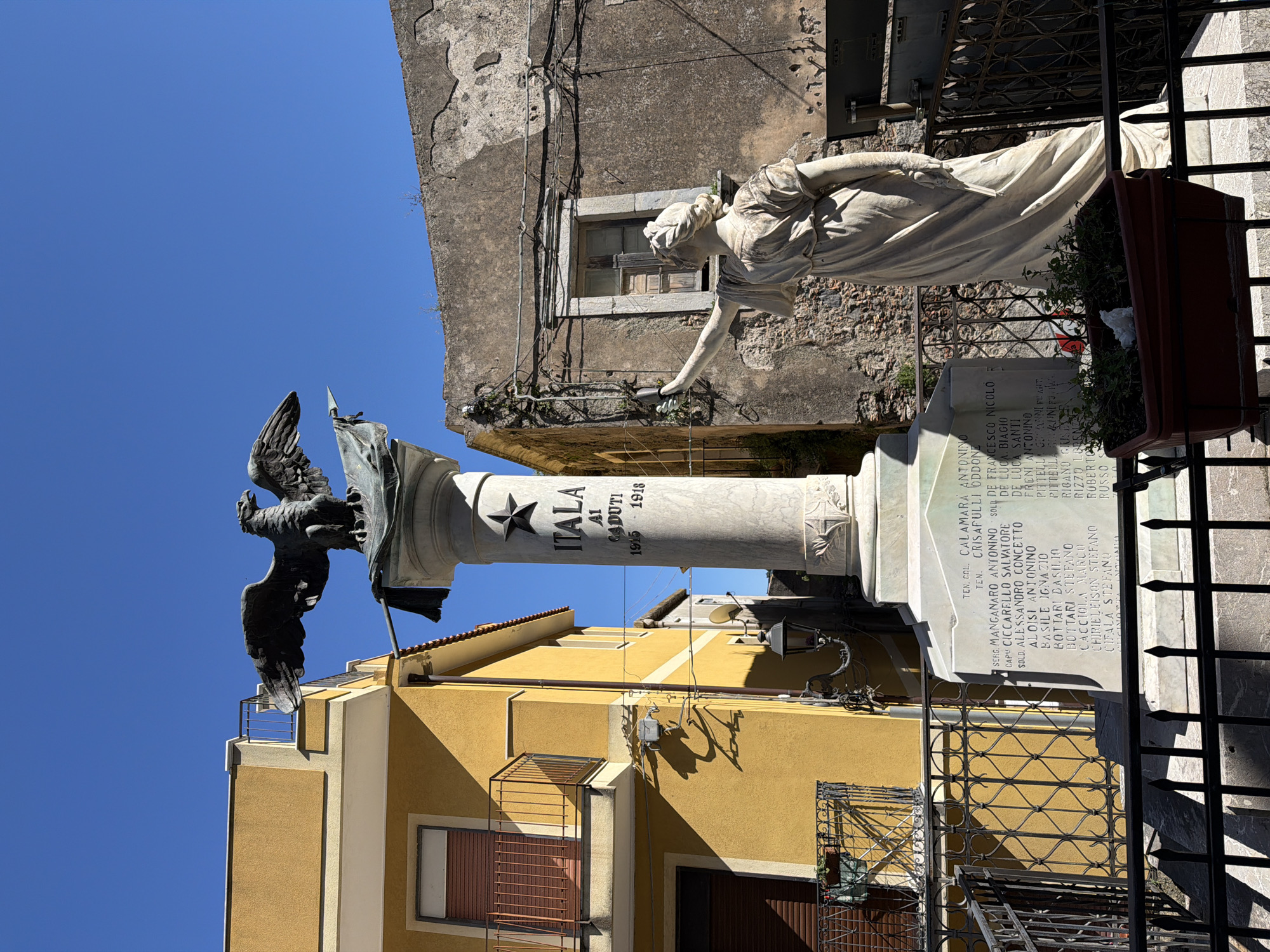 A war memorial stands prominently in a bright, sunlit outdoor setting, likely a town square or street. The central feature is a tall, light-colored column topped by a dark, metallic eagle with outstretched wings, holding what appears to be a flag or standard. Below the eagle, the column bears the inscription "ITALA Al CADUTI 1915 - 1918," indicating it is a memorial to those who fell during World War I, specifically from Itala, Italy.
To the right of the column (when viewed upright), a graceful white marble statue of a female figure is positioned, seemingly reaching out or gesturing towards the memorial. The statue is partially obscured by a dark metal fence and a planter box filled with green foliage. Below the statue, a base or plinth lists numerous names, likely the fallen, including ranks such as "TEN. COL.," "TEN.," "SERG.," "CAP.," and "SOLD." Specific names visible include "CALAMARA ANTONINO," "CRISAFULLI ODDONE," "MANGANARO ANTONINO," "CICCARELLO SALVATORE," "ALESSANDRO CONCETTO," "ALOISI ANTONINO," "DE LUCA BIAGIO," "DE LUCA SANTI," "FRENI ANTONINO," "PITTELLA GIANNI," "BASILE IGNAZIO," "BOTTARI BASILIO," "BOTTARI STEFANO," "CACCIOLA MARCO," "CHIRIELEISON STEFANO," and "CIFALA STEFANO."
The memorial is flanked by buildings: an old, weathered grey building with a window behind the female statue, and a vibrant yellow building with a tiled roof, visible behind the column and eagle. The clear blue sky and strong shadows suggest bright daylight, indicating good weather. The overall scene conveys a sense of solemn remembrance in a historic European town context.