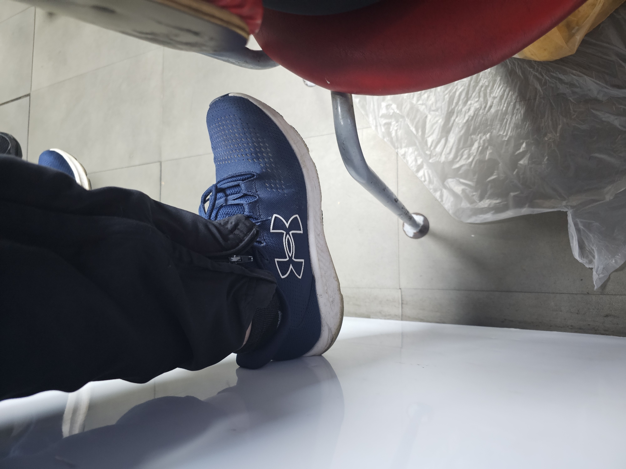 A person wearing blue Under Armour sneakers and dark blue pants is seated on a red chair with metal legs. The chair is positioned on a white tiled floor. The background includes a partially visible second shoe, what appears to be a plastic bag or wrap, and sections of grey tiled flooring. There is no verifiable information about Holywell, United Kingdom in the image.