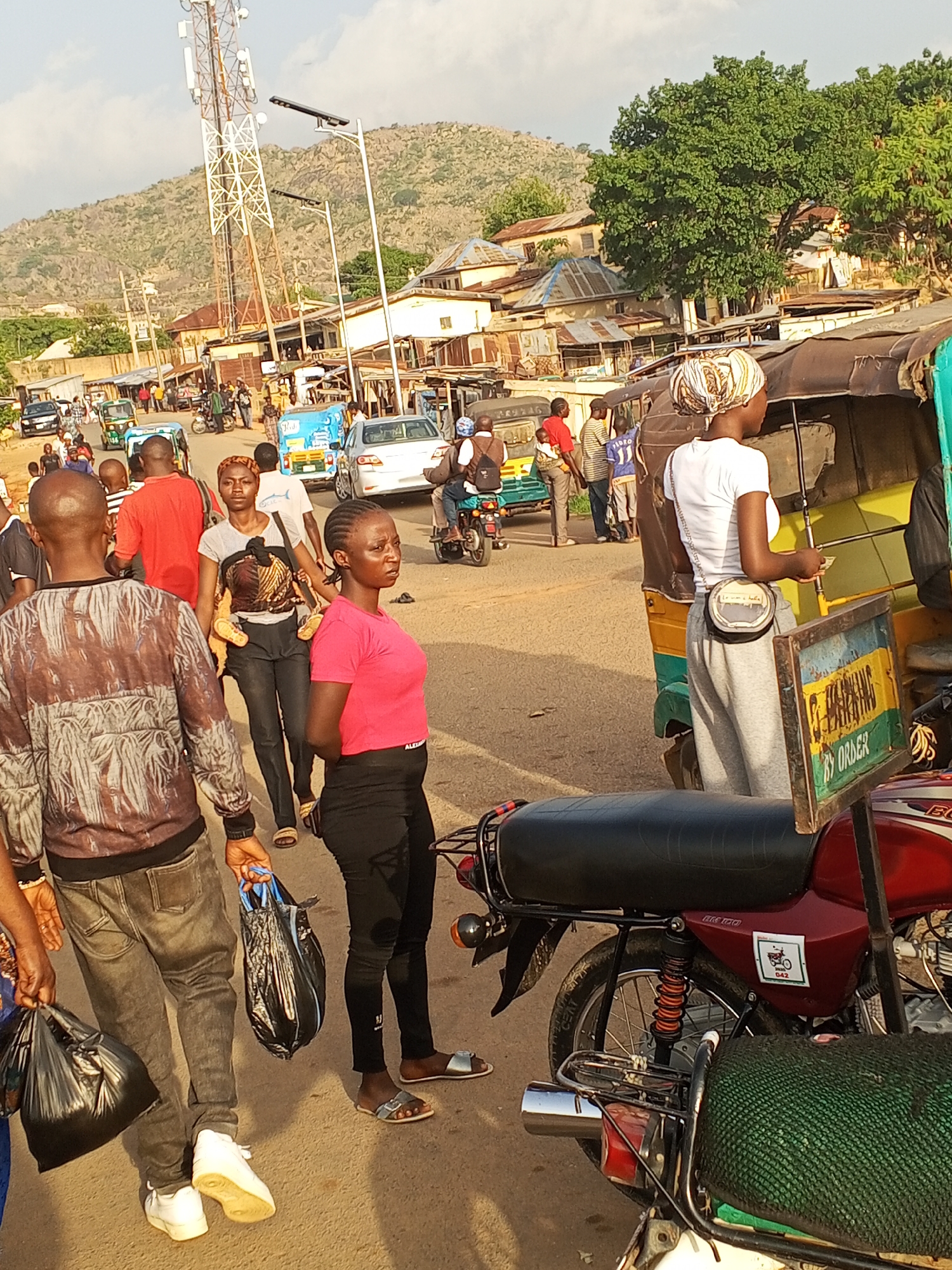 The image depicts a street scene in Gwong, Nigeria. A mix of pedestrians and vehicles, including motorcycles and auto-rickshaws (tuk-tuks), occupy the street.
Several people are visible, including a woman in a pink shirt and black leggings, a woman in a white shirt and gray sweatpants, and a man in a patterned sweater and jeans. They appear to be waiting or walking along the roadside. Other individuals are dispersed throughout the scene, some interacting with vehicles.
The background includes buildings with corrugated metal roofs and a telecommunications tower against a hilly landscape. Overhead utility wires are visible. The general environment suggests a populated, developing urban area.