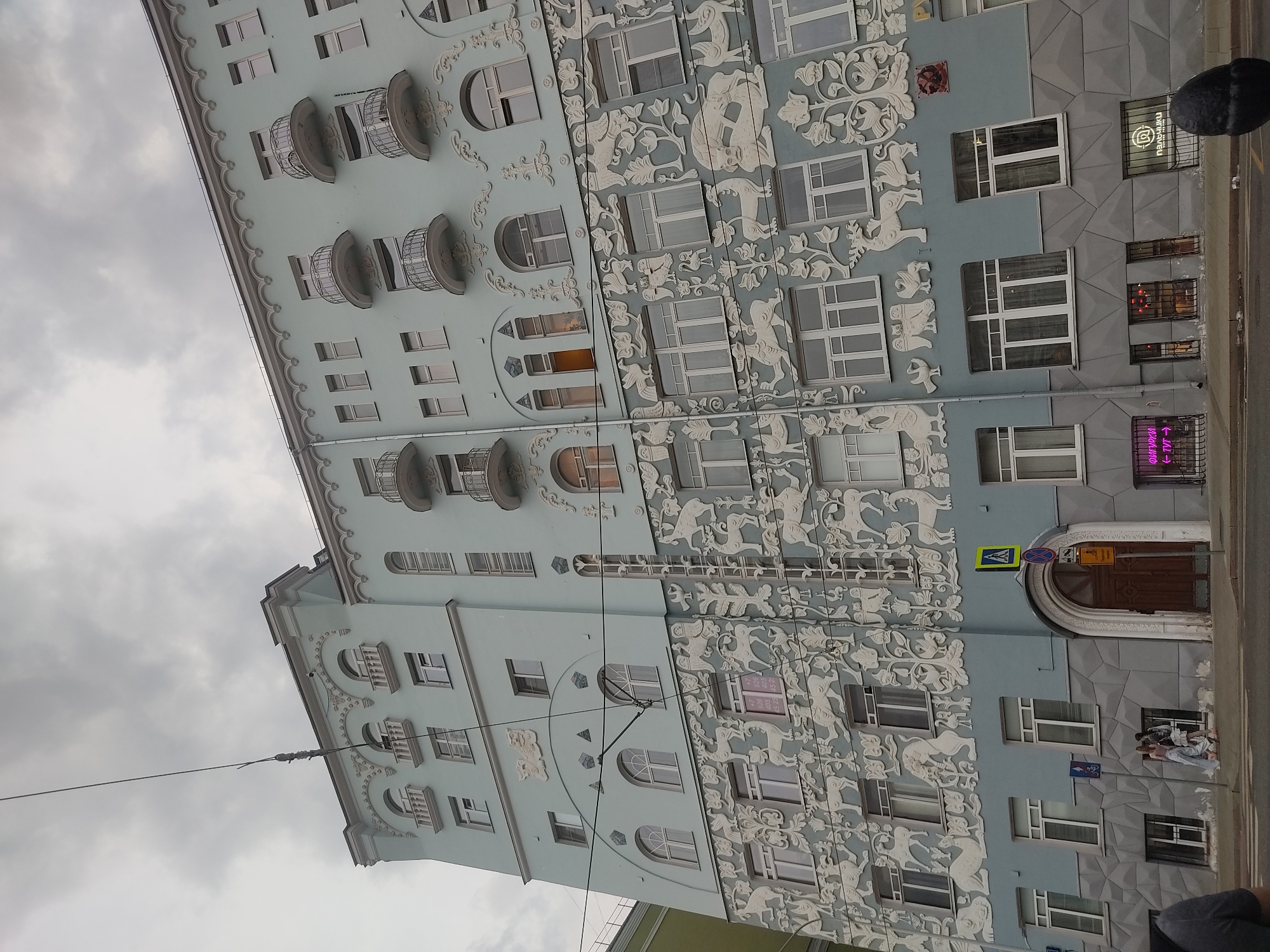 The image depicts a building in Moscow, Russia, likely located in the city center, constructed in Art Nouveau style. The building features a light blue facade, embellished with stucco reliefs of fantastical creatures and floral motifs. It has multiple windows, some of which are adorned with ornate balconies.
On the right-hand side of the building, the facade transitions to a grey, textured finish with less ornate features. Signage and window displays indicate commercial activity at street level. Pedestrians and street infrastructure, such as road signs, are visible in the foreground. The sky above is overcast with grey clouds.