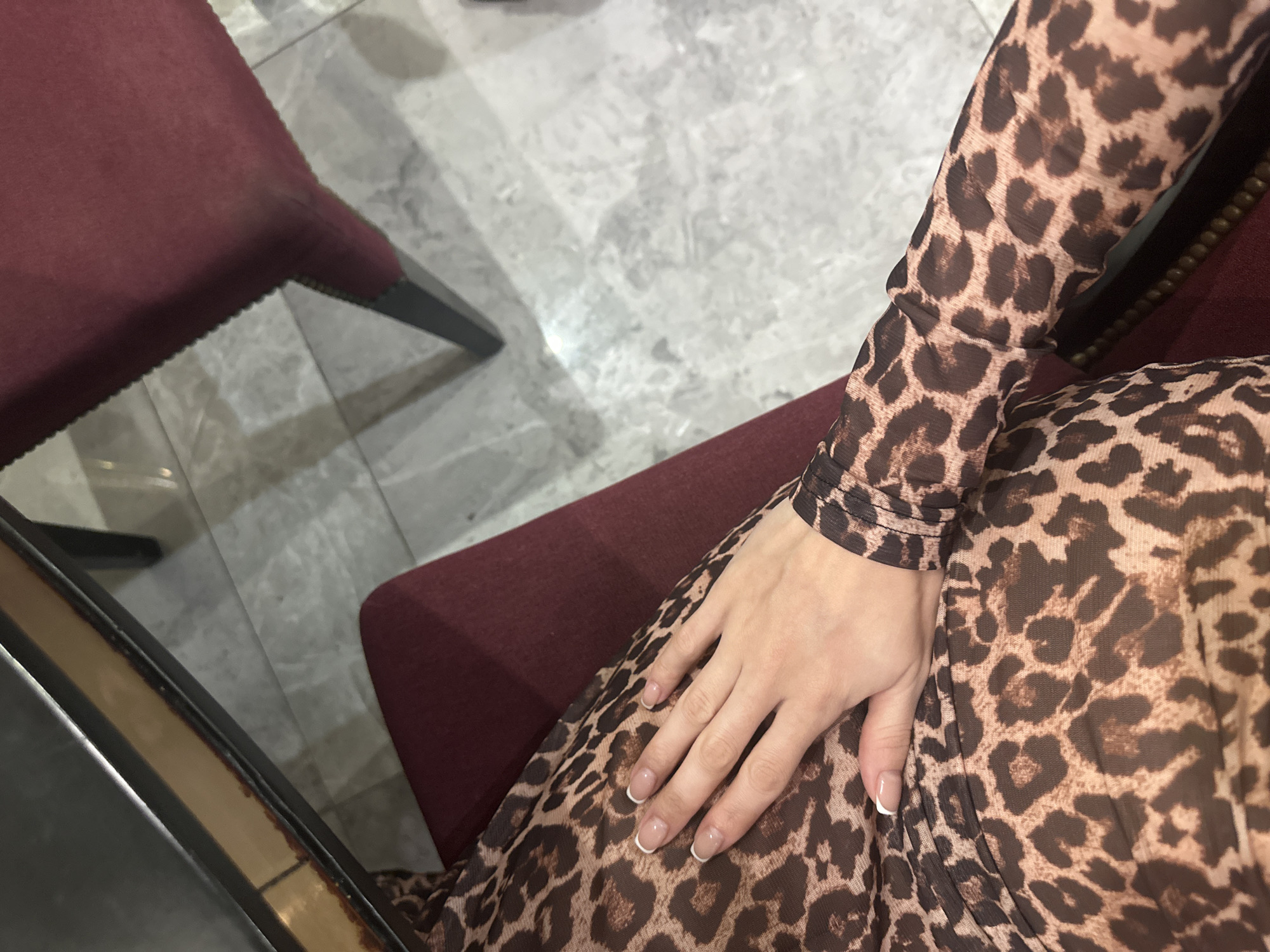 Here is a detailed description of the image:
The image shows a person seated, wearing a long-sleeved leopard print garment. The person's hand is resting on the garment, displaying a French manicure. The person is sitting on a red upholstered chair with dark wooden legs. Another red chair is partially visible on the left. The background consists of a gray and white marble tile floor. The location is Unknown, Unknown.