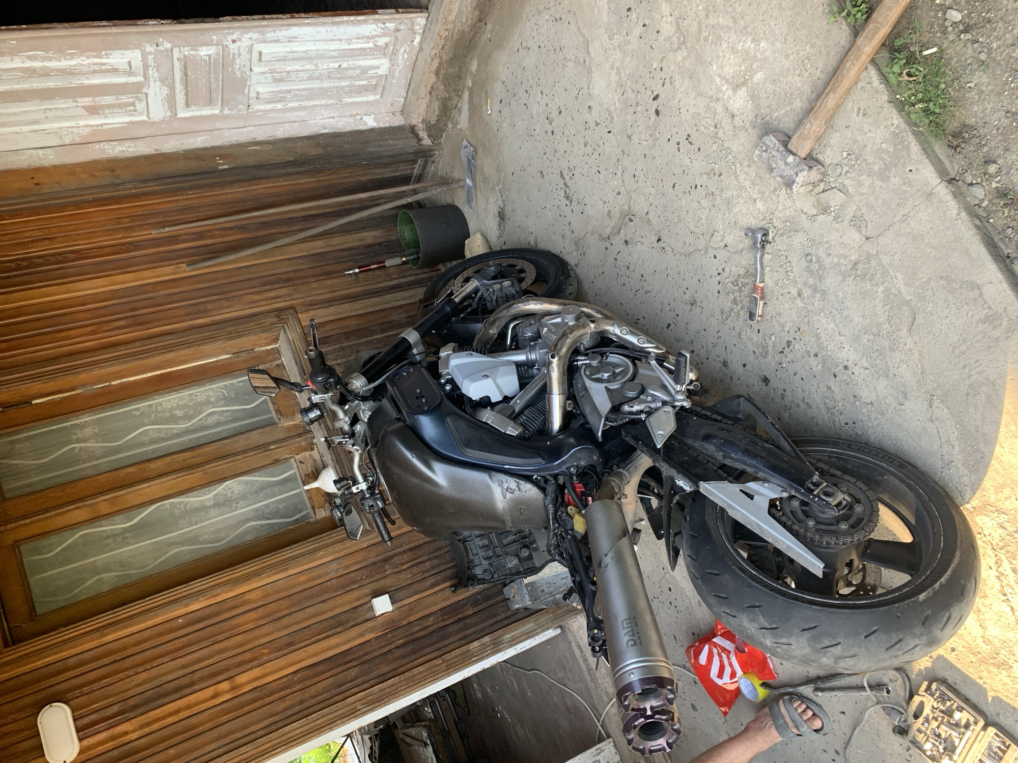 A motorcycle, partially disassembled, sits on a concrete surface in front of a weathered wooden building with large, paneled doors and windows. The motorcycle is dark-colored with a prominent exhaust pipe and various mechanical components exposed. Tools, including a socket wrench set and a sledgehammer, are scattered on the ground near the motorcycle. A person's bare foot, wearing a sandal, is visible on the left side of the frame. The scene appears to be an outdoor workspace, possibly a garage or yard. The environment suggests a rural or semi-rural setting, with vegetation visible in the background.