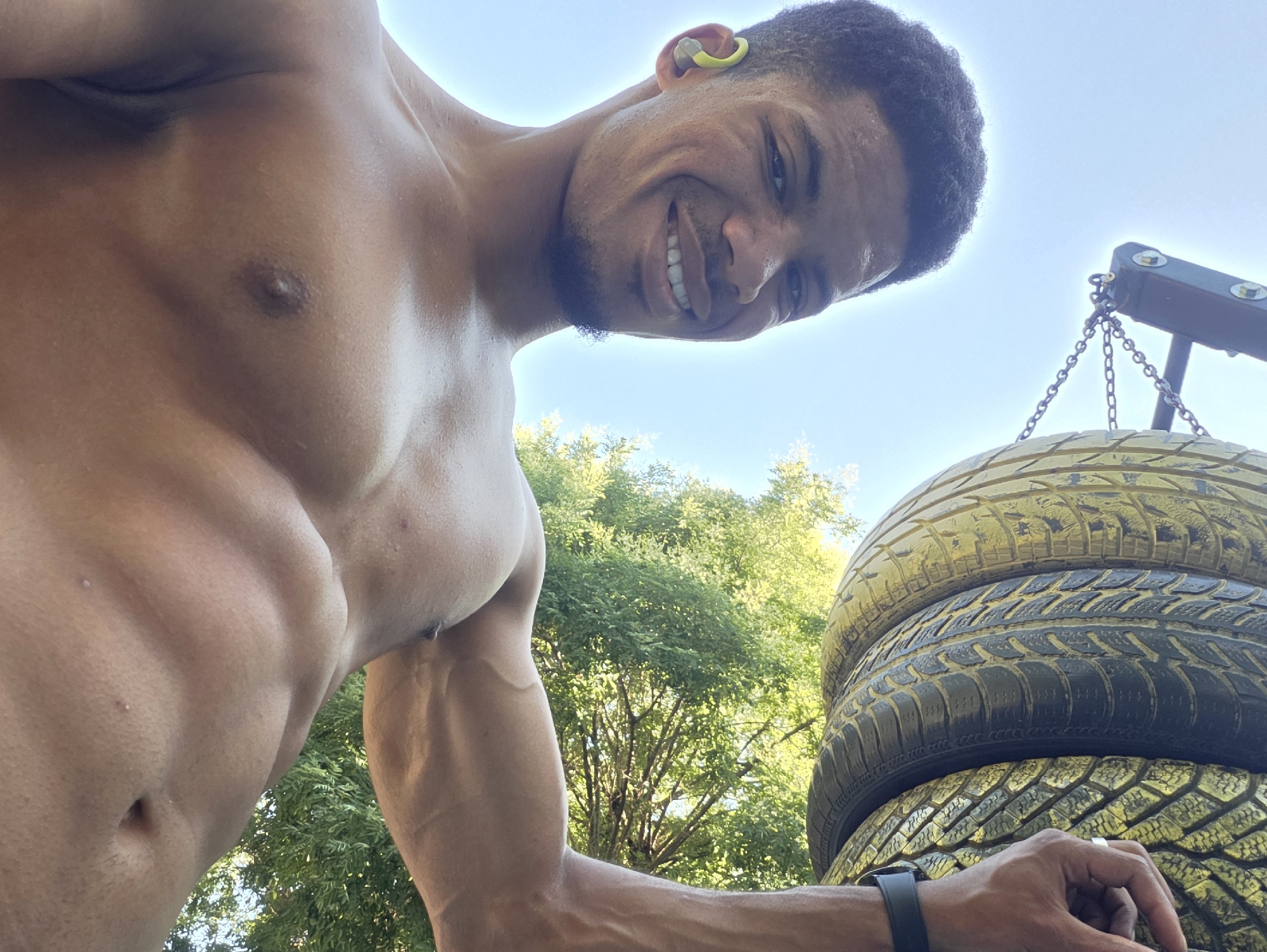 A low-angle, close-up shot shows a shirtless, muscular man outdoors, with a bright blue sky and green trees in the background. The man has short, dark, curly hair and is wearing a yellow earbud in his right ear. He is smiling and looking down at the camera. His chest and abdomen are visible, revealing well-defined muscles. To the right of the man, a stack of four tires is visible, suspended by chains from a dark wooden structure. The tires appear to be old and weathered, with some painted yellow. The man's right arm is extended towards the tires, and his hand is resting on the bottom tire. A black smartwatch is visible on his left wrist. The lighting suggests it is a sunny day. The specific location in the city of Varna, Bulgaria cannot be determined from the visual information provided.