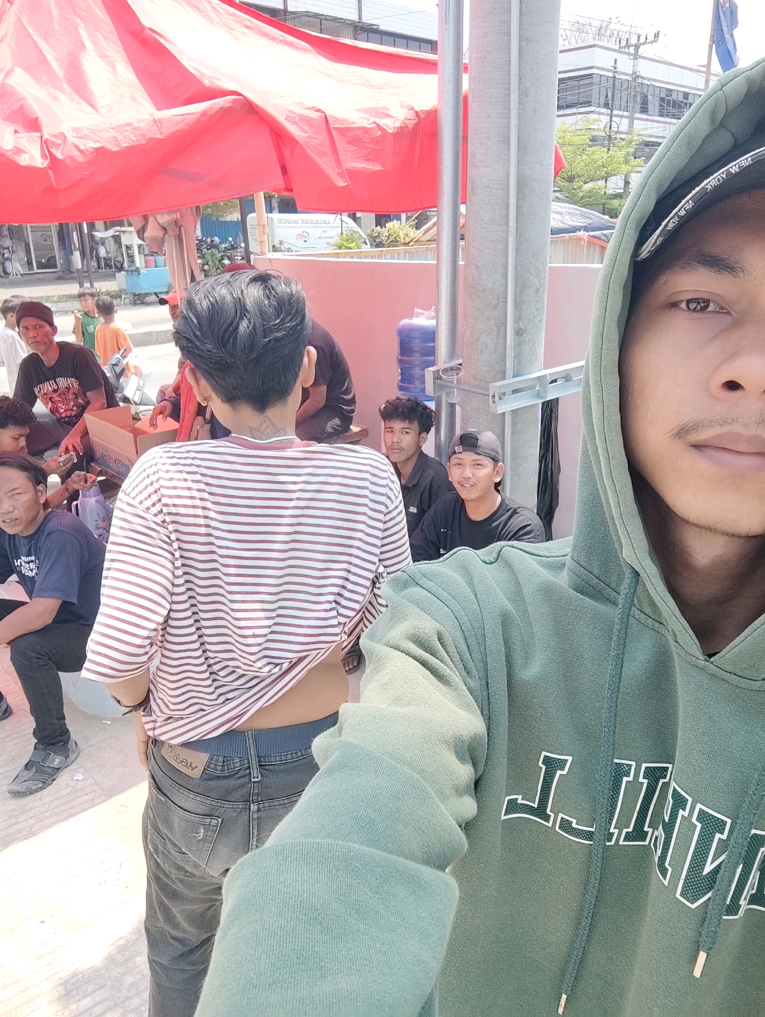 A group of young men are gathered outdoors, with a large red canopy tent providing shade. One man is in the foreground, taking a selfie with his right arm extended. He is wearing a green hooded sweatshirt and a black baseball cap. Behind him, several other men are visible. One man in a striped shirt is facing away from the camera, his back to the viewer. Other men are sitting on the ground or on a low wall. There are boxes and a water cooler in the background, suggesting a casual gathering or a small market stall. The environment appears to be a public space with some buildings and utility poles visible.