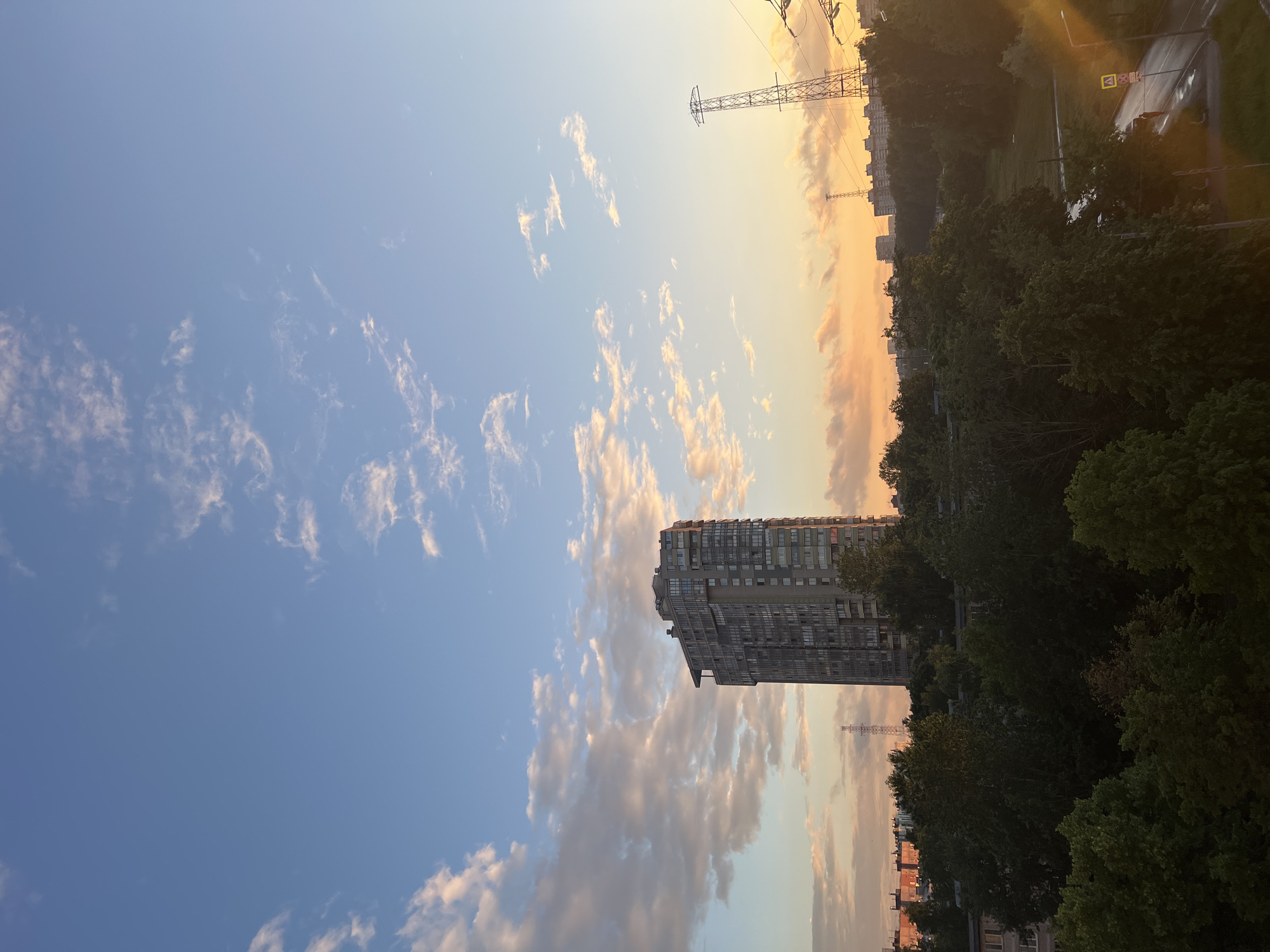 The image depicts a city skyline at sunset, with a focus on the sky and the urban landscape. Tall, grey apartment buildings with many windows dominate the foreground on the left, contrasted by a dense line of green trees along the right side of the frame. In the distance, more buildings and a large, lattice-like transmission tower are visible.
The sky is a gradient of colors, transitioning from a clear, light blue at the top to warm hues of orange and yellow near the horizon, suggesting the sun is setting. Scattered, wispy clouds catch the light, appearing pink and gold. The overall impression is one of an urban environment bathed in the soft, warm light of dusk.
The scene appears to be viewed from a high vantage point, possibly a window or balcony. The presence of residential buildings, trees, and a transmission tower indicates an urban setting, likely within a city. No people are visible in the image. The weather appears clear, with the sunset indicating the time of day. There are no discernible signs or text in the image.