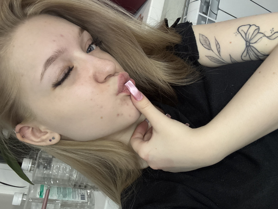 The image displays a close-up portrait of a young female with light skin and blonde or light brown hair, posing indoors. She is wearing a dark, likely black, short-sleeved garment. Her left eye is partially closed in a wink, revealing a blue iris and long eyelashes. Her lips are pursed in a "kissing" expression, with her left index finger touching them. The fingernails on her visible hand are long, square-tipped, and coated in a light pink or clear finish. Her left ear exhibits multiple piercings, including a helix hoop and two lobe studs. Several blemishes are visible on her facial skin. On her upper left arm, a tattoo depicting a leafy branch and a ribbon or bow design is visible. The background to the left shows several clear plastic bottles, some containing liquid, and a green plant leaf. To the right, a white, textured object, possibly a radiator or screen, and a reddish-brown vertical surface are partially visible. The specific location within Saratov, Russia, cannot be determined from the provided visual information.