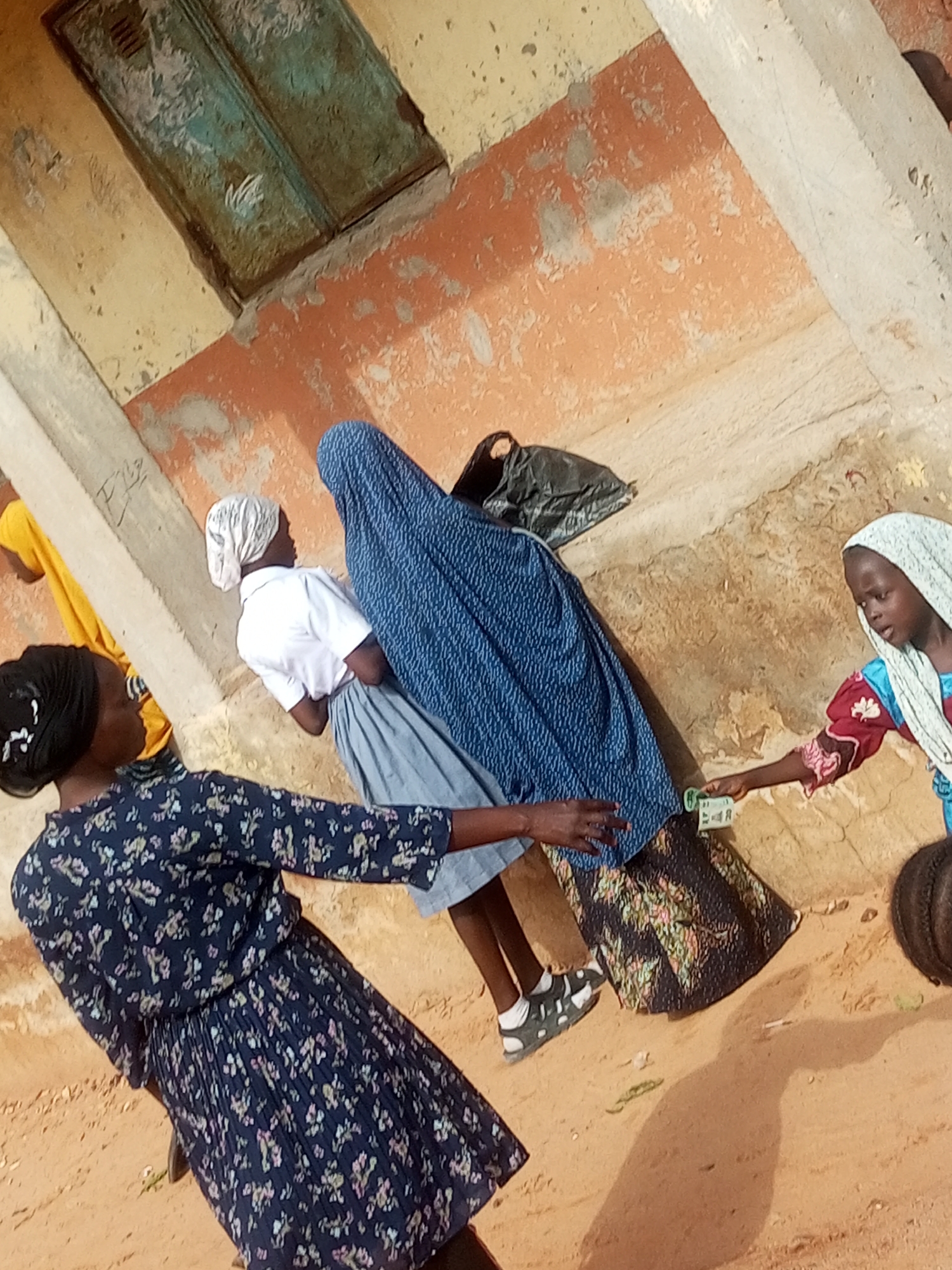 The image shows a street scene in Tempure, Nigeria, with several individuals. In the foreground, a person wearing a dark blue floral dress and a black headscarf is walking away from the camera. To their right, a young girl in a white headscarf and a white shirt with a blue skirt is being handed some money by another person. A large blue garment is draped over something behind the young girl. Further back, a person in a yellow garment is partially visible. The ground is sandy, and there is a textured wall behind the individuals. The overall impression is a candid snapshot of daily life in a community.
